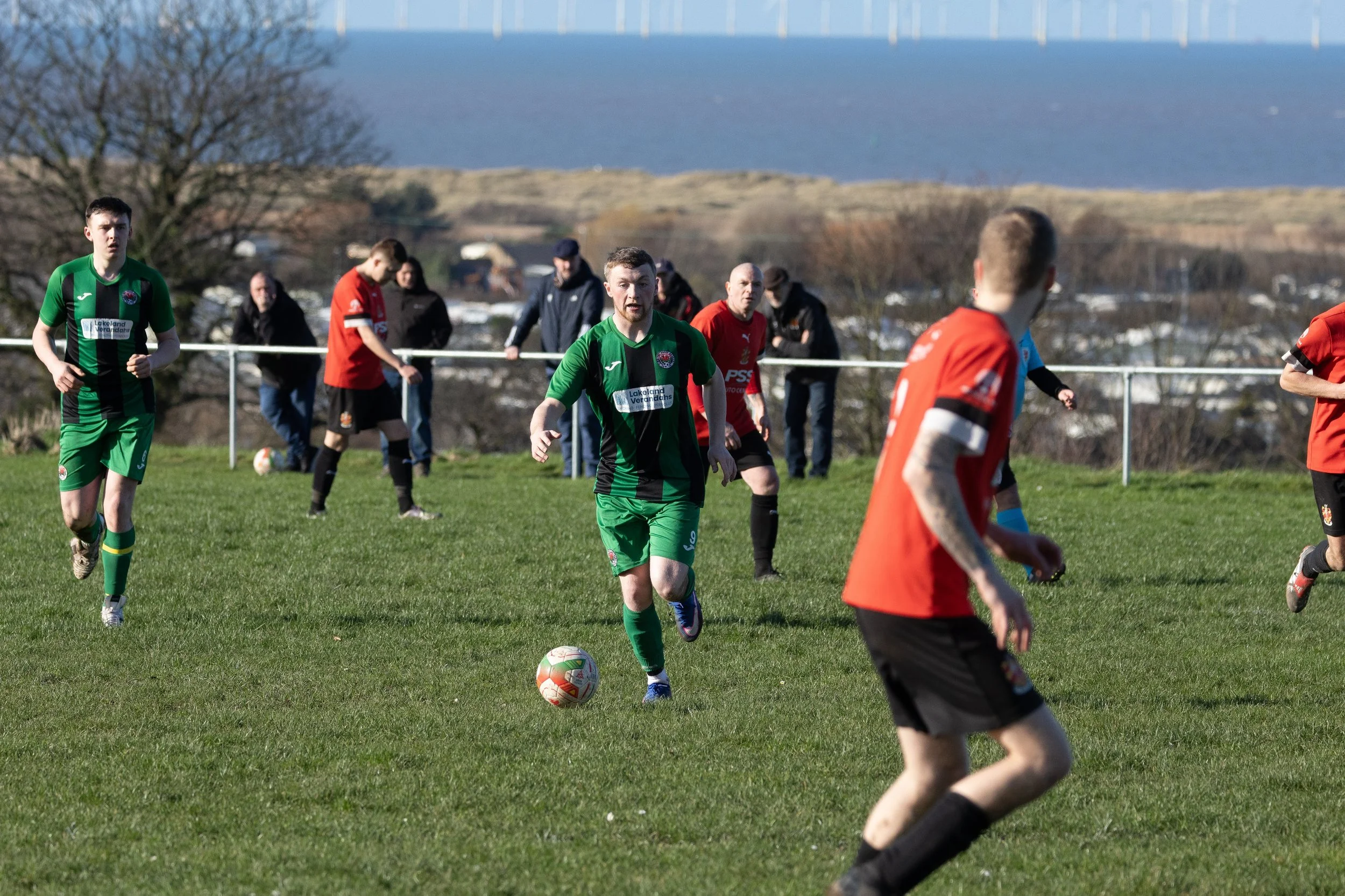 Soccer players in green and red uniforms on a grassy field with a scenic landscape background.