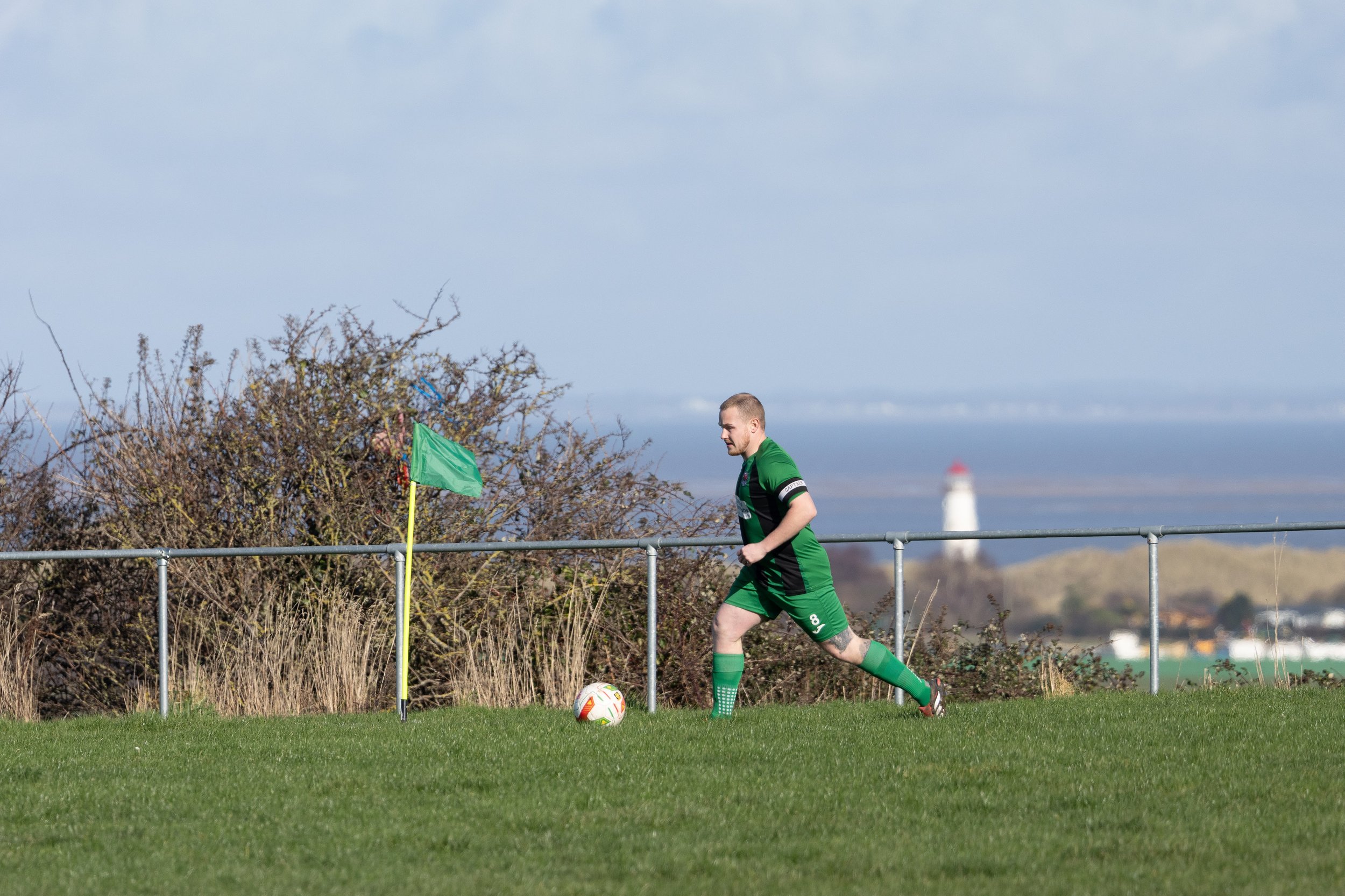 A man in a green soccer uniform is running on a grassy field near a metal fence, with a soccer ball near his feet, and a lighthouse in the background on a cloudy day.