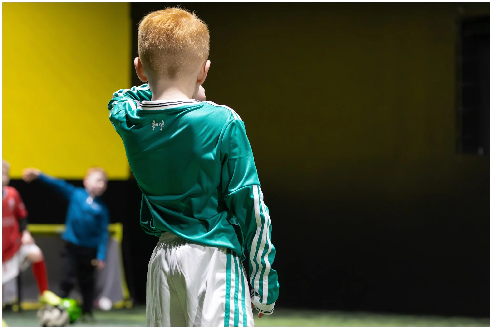 A young boy with red hair wearing a teal sports jersey and white shorts, standing on a soccer field, with other kids in sportswear in the background under a yellow and black wall.
