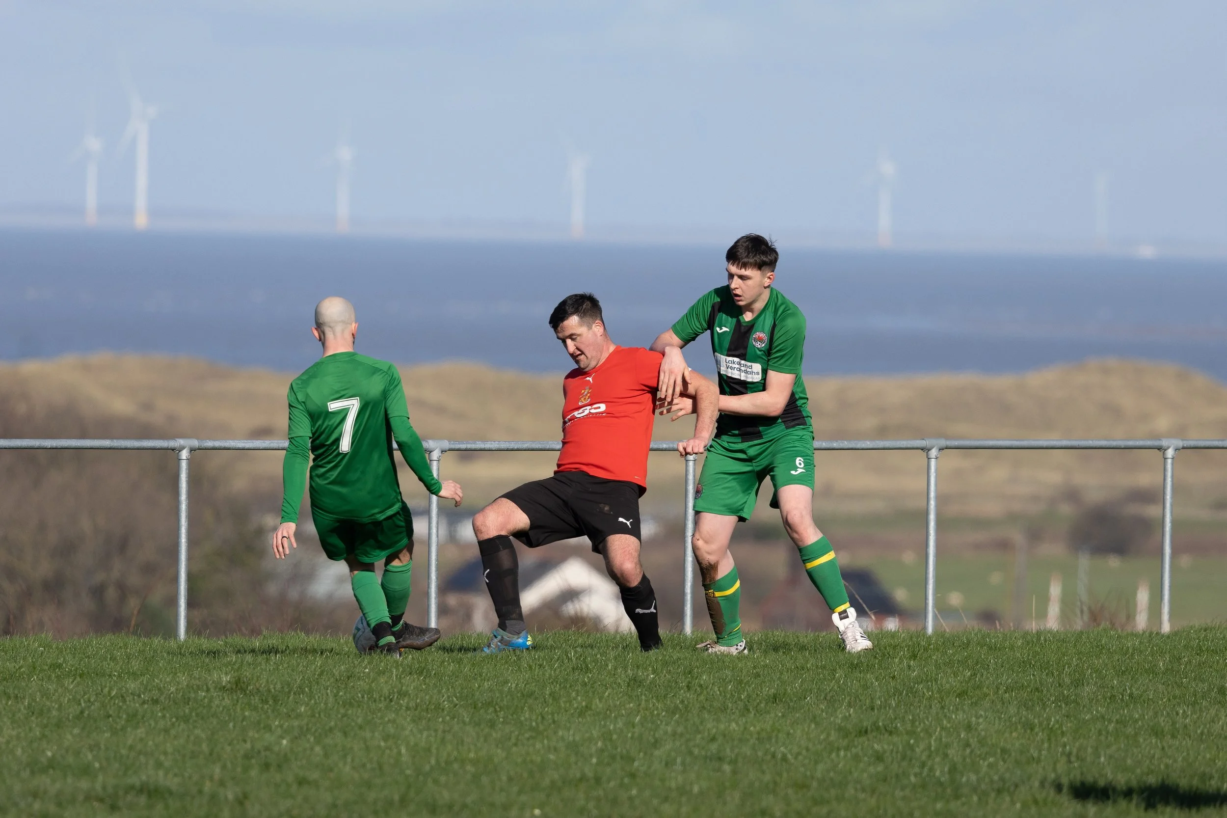 Soccer players in green and red jerseys struggle for ball control near the sideline of a field with wind turbines in the background.
