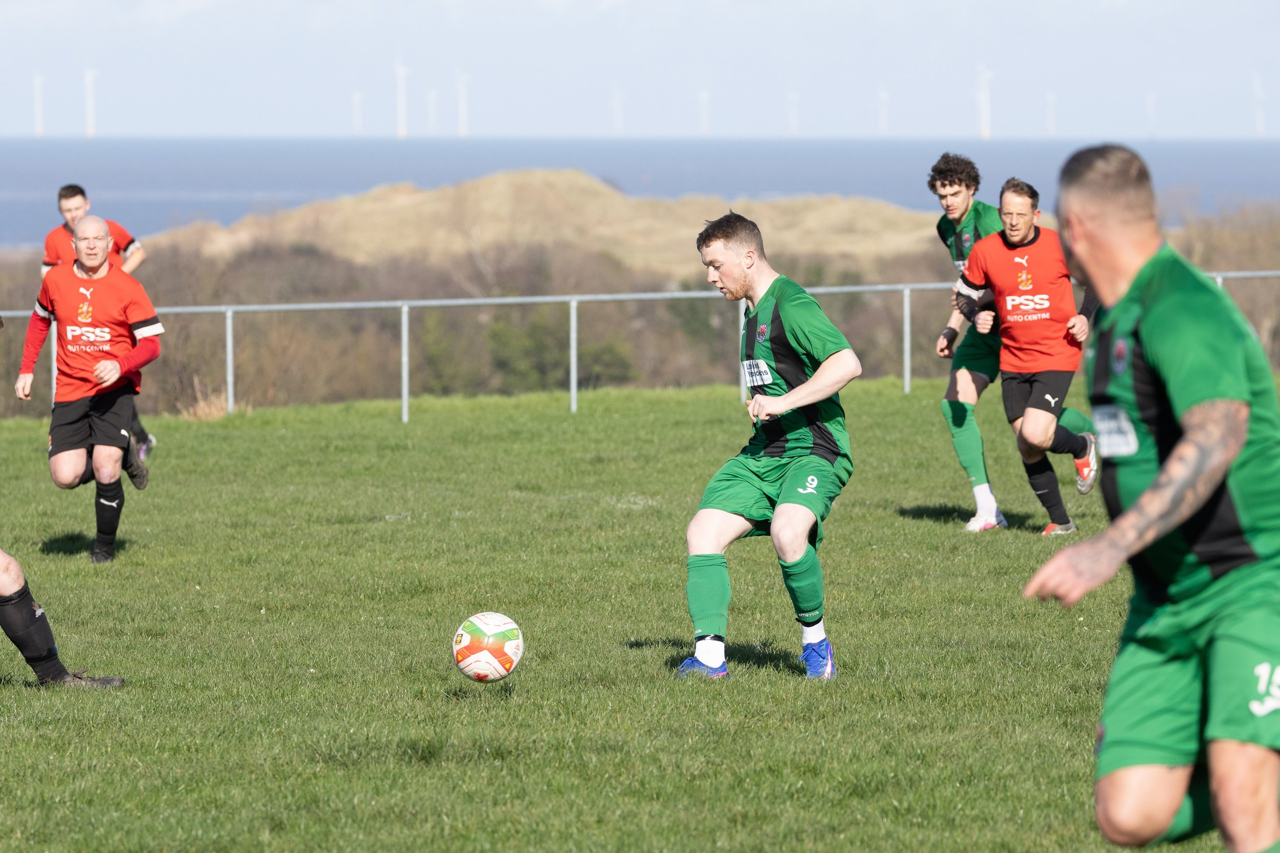Soccer players on a grassy field, one in green about to kick the ball, others around him, with hills and wind turbines in the background.