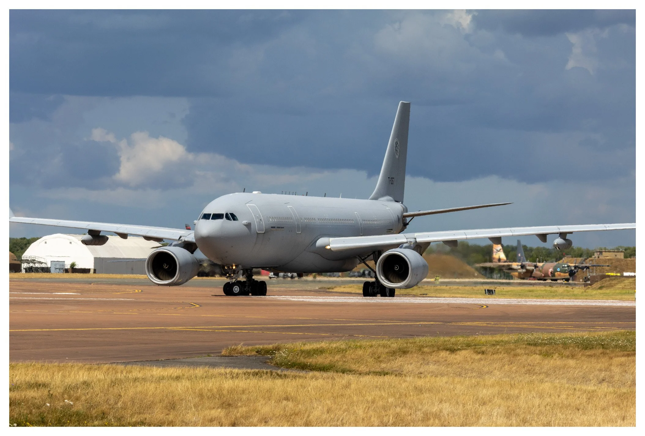 A large military aircraft taxiing on an airport runway with a dark cloudy sky overhead and several other aircraft in the background.