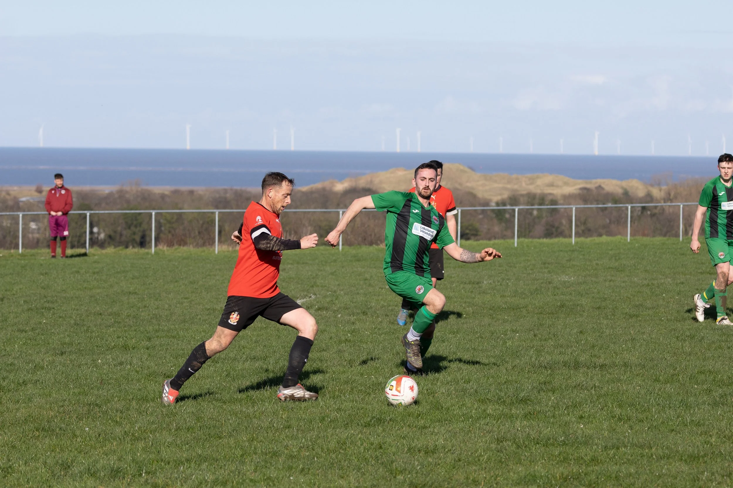 Soccer match on grassy field with players in red and green uniforms, near a fence with ocean and wind turbines in the background.