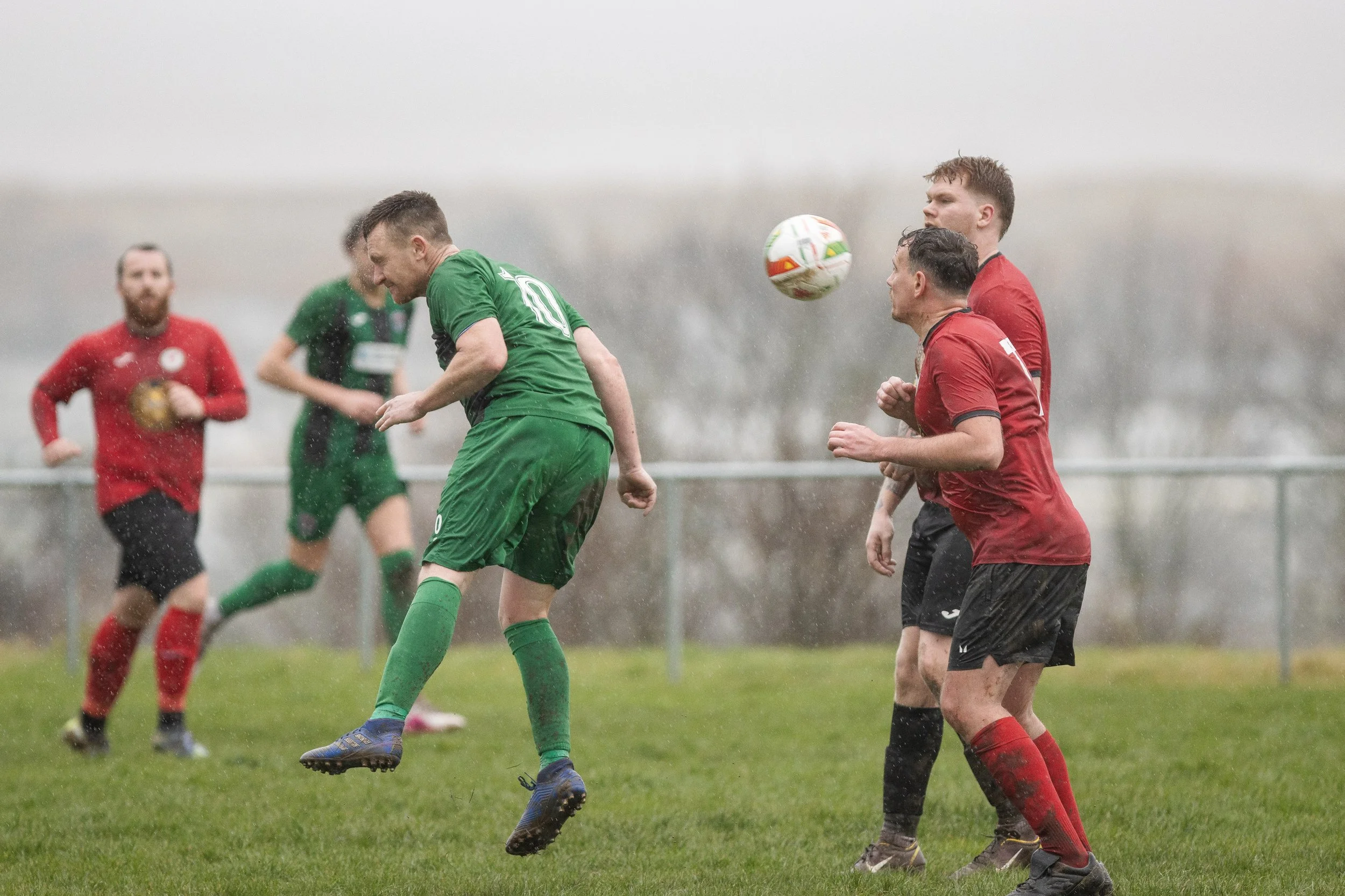 Soccer match in the rain with players in green and red jerseys competing for the ball on a grassy field.