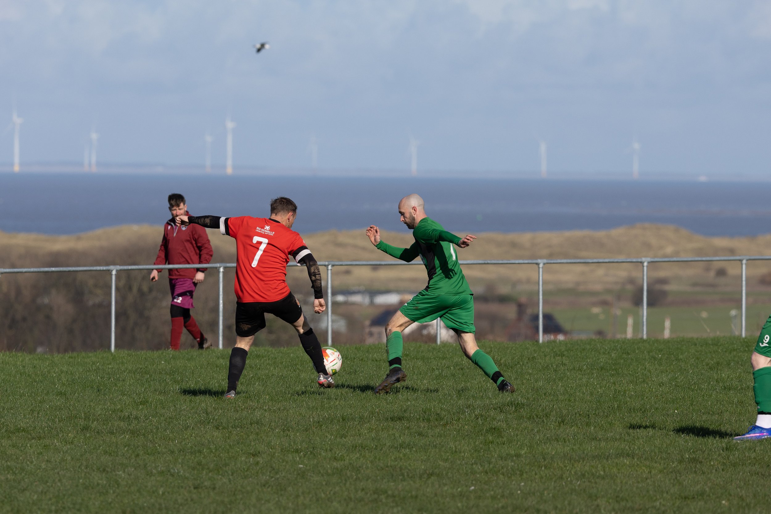Two soccer players, one in a red jersey with the number 7, and the other in a green jersey, are competing for the ball on a grassy field. A third person in the background is wearing a maroon jacket and maroon pants, and is watching the game. Wind tur