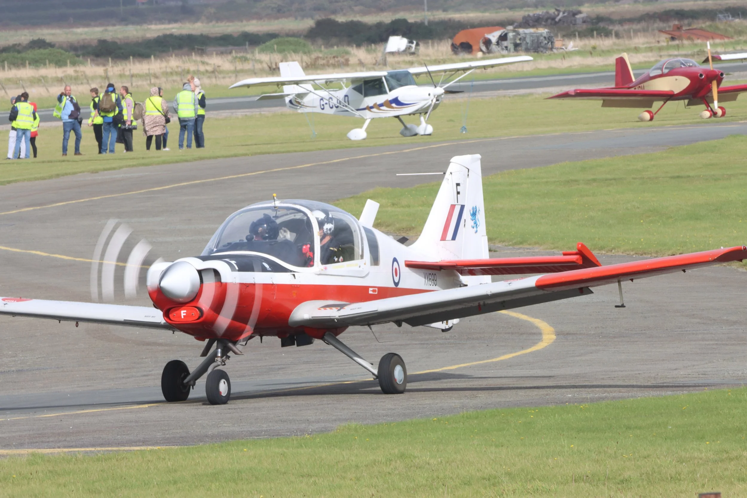 A red and white military jet taxiing on the runway at an airport during an airshow, with a group of people in high-visibility vests and other small aircraft in the background.