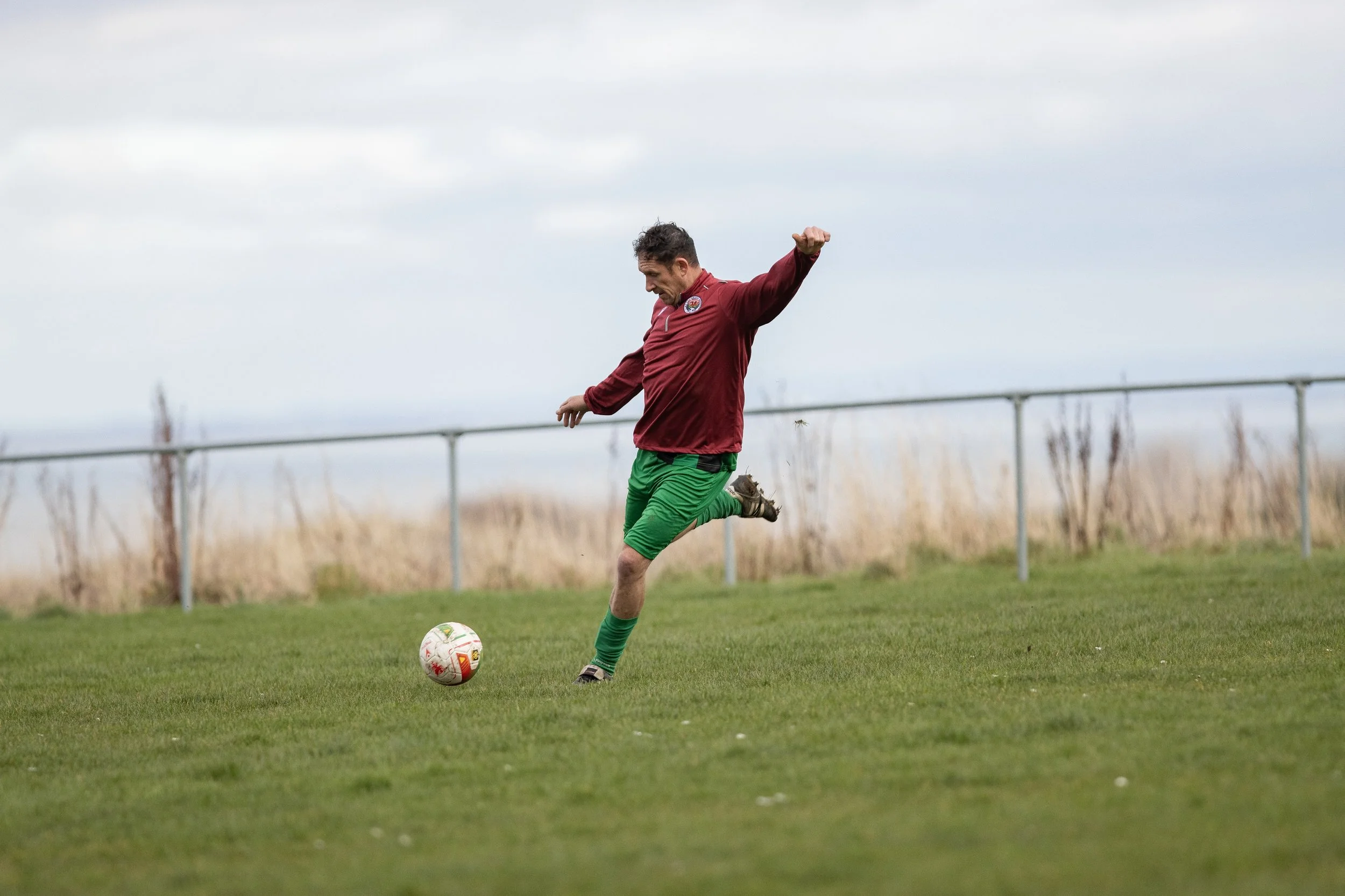 A man kicking a soccer ball on a grassy field with a cloudy sky and a fence in the background.
