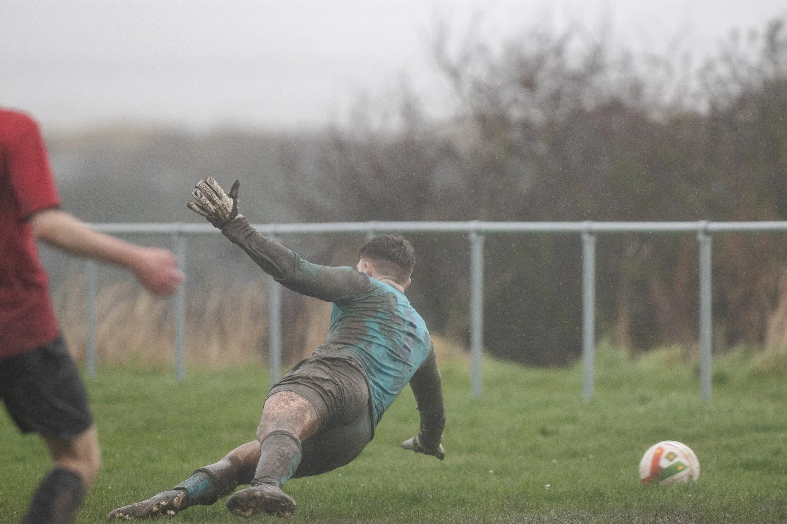 A soccer goalkeeper dives to block the ball during a match on a rainy day, with a player in a red jersey partially visible in the foreground.