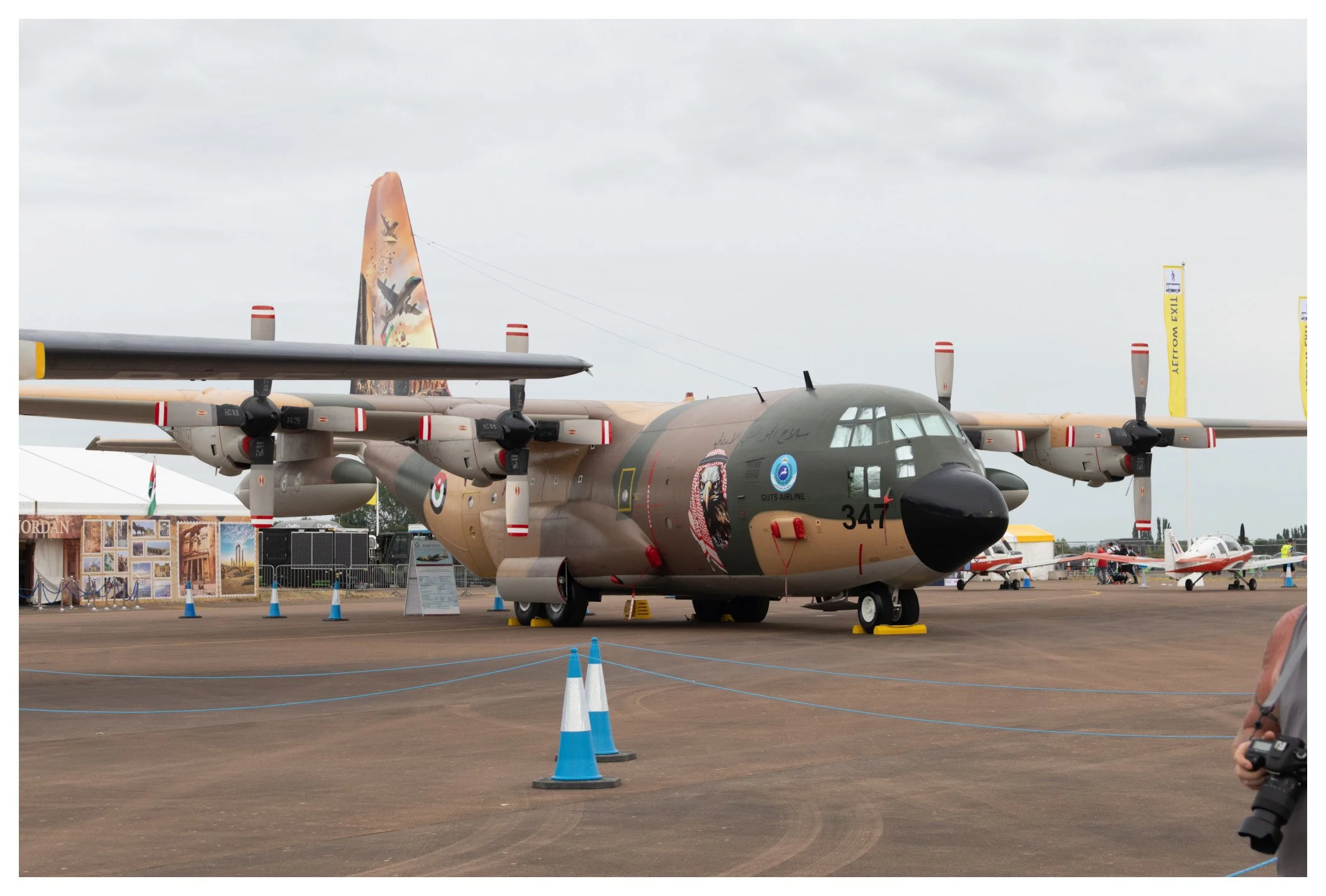 Military aircraft on display at an air show, painted with camouflage and military markings, with other small planes in the background.