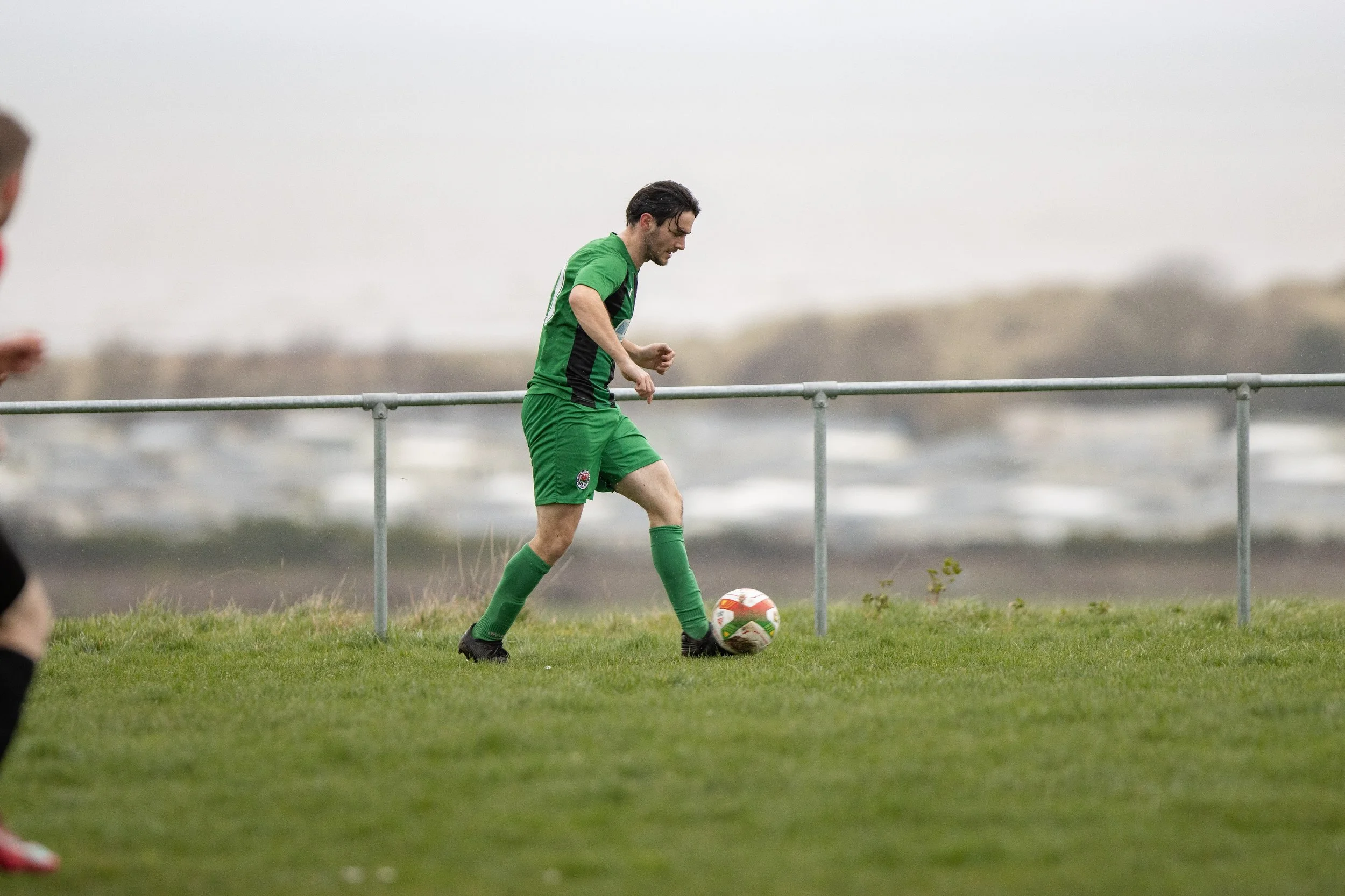 A soccer player in a green uniform preparing to kick a soccer ball on a grassy field during an overcast day.