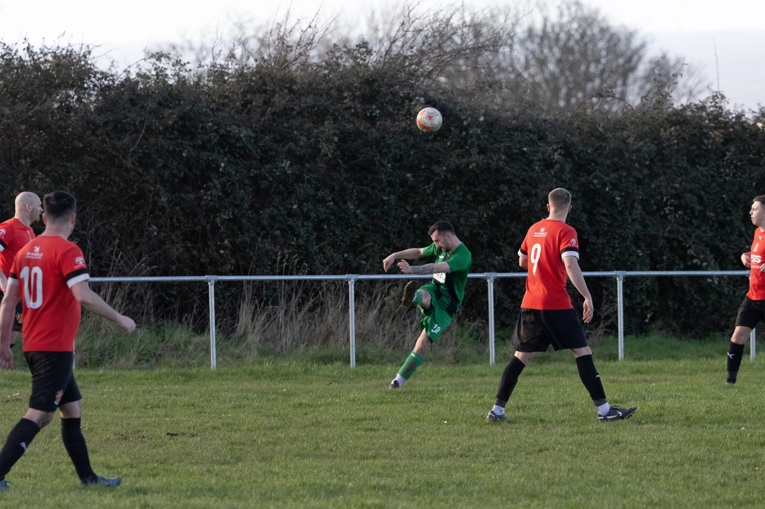 Soccer players in red and green uniforms playing on a grassy field, with the ball in mid-air against a background of dark bushes and gray sky.