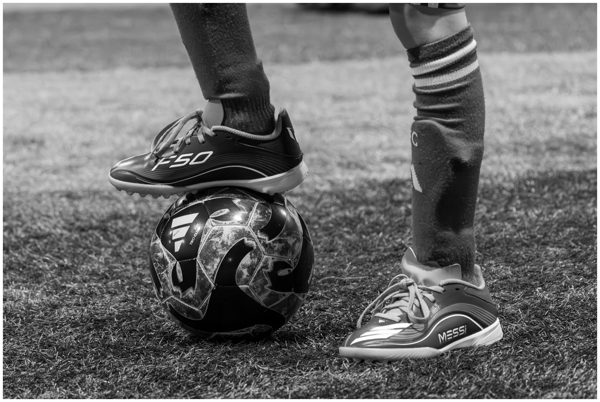 Close-up of a soccer player's feet, with one foot wearing a firm athletic shoe on top of a patterned soccer ball, on grass field. The player is wearing striped socks and athletic pants.