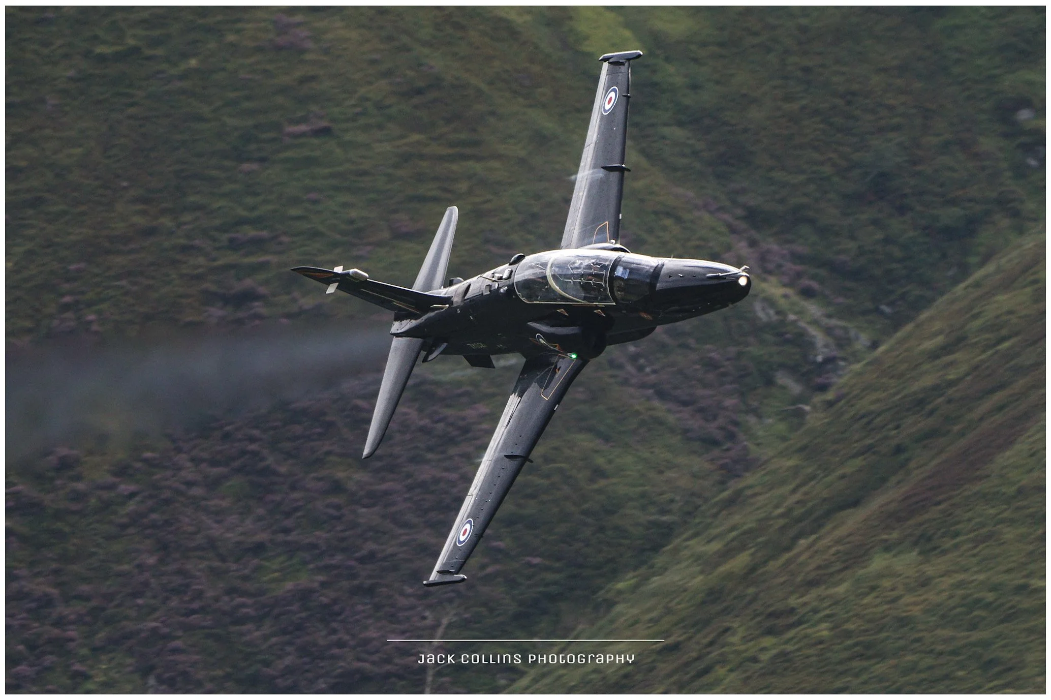 A sleek black jet aircraft flying at an angle over green mountainous terrain.