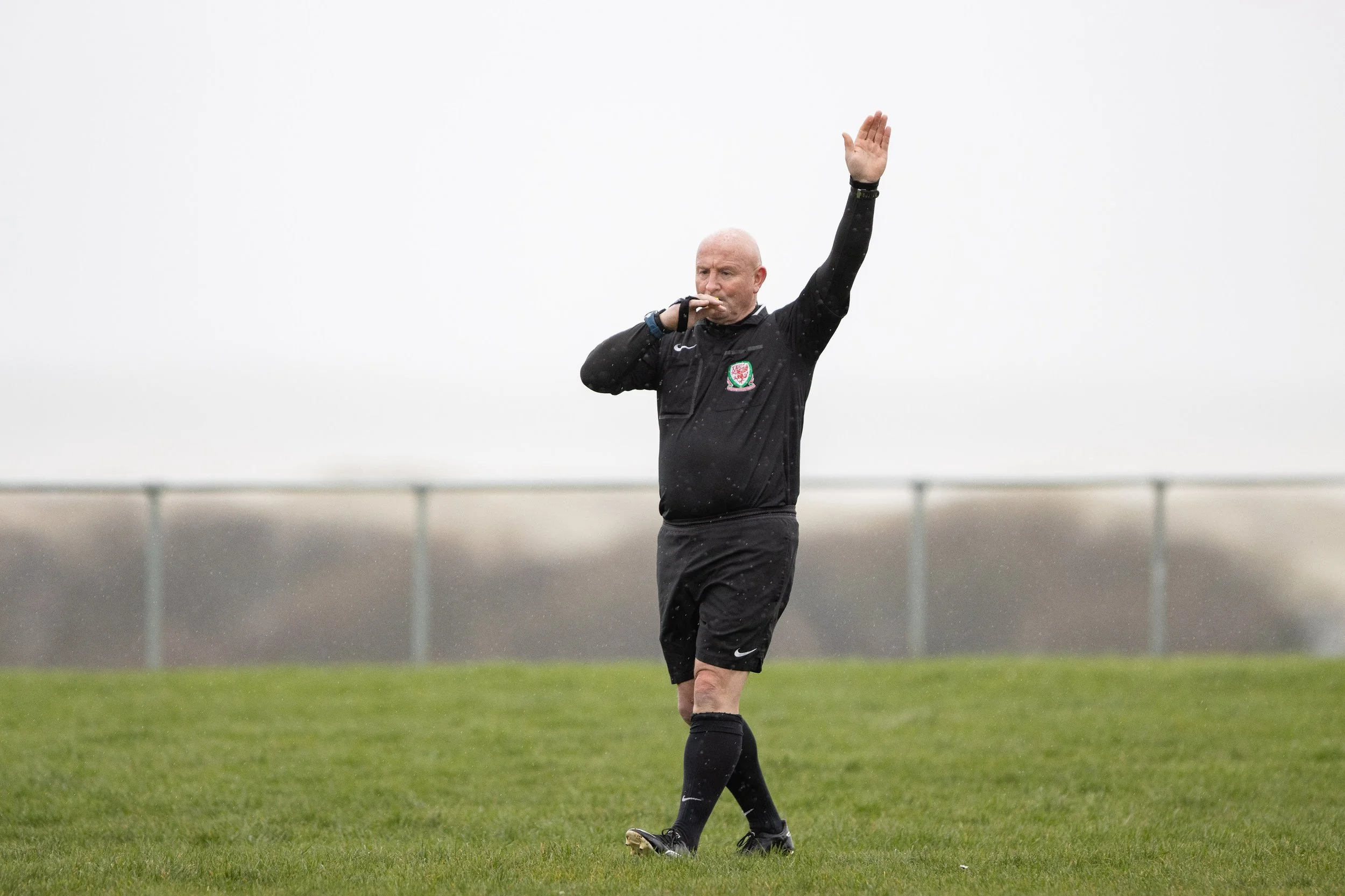 A soccer referee in black uniform making a call during a game, standing on grass with a fence in the background.