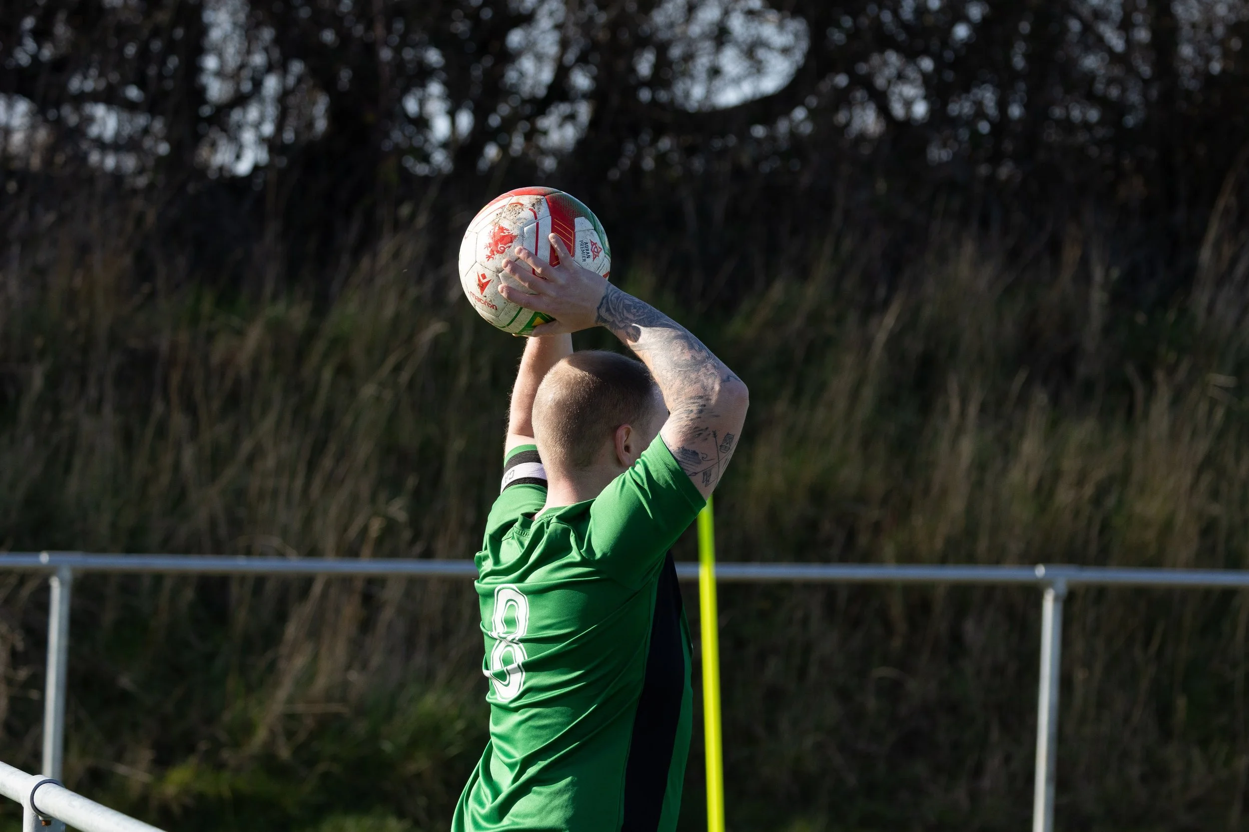 A soccer player wearing a green jersey with the number 8 practices throwing a soccer ball over his head outdoors during daylight, with a background of trees and grass.