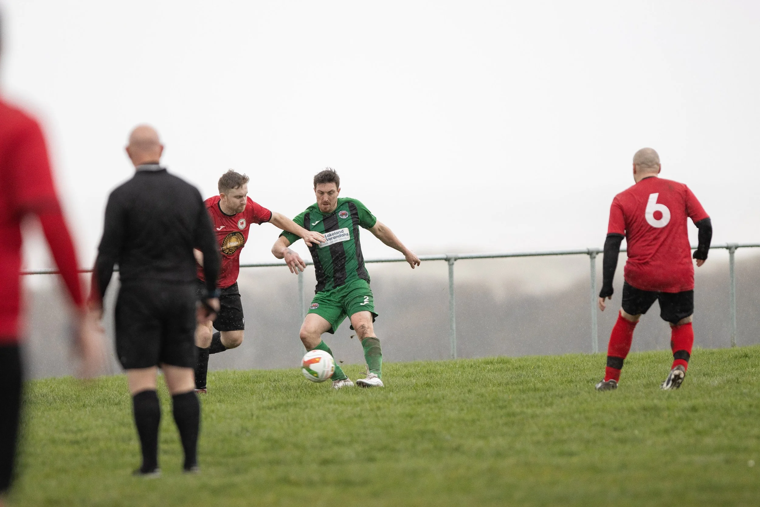Soccer match with players competing for the ball on the field under cloudy weather, with a metal fence in the background.