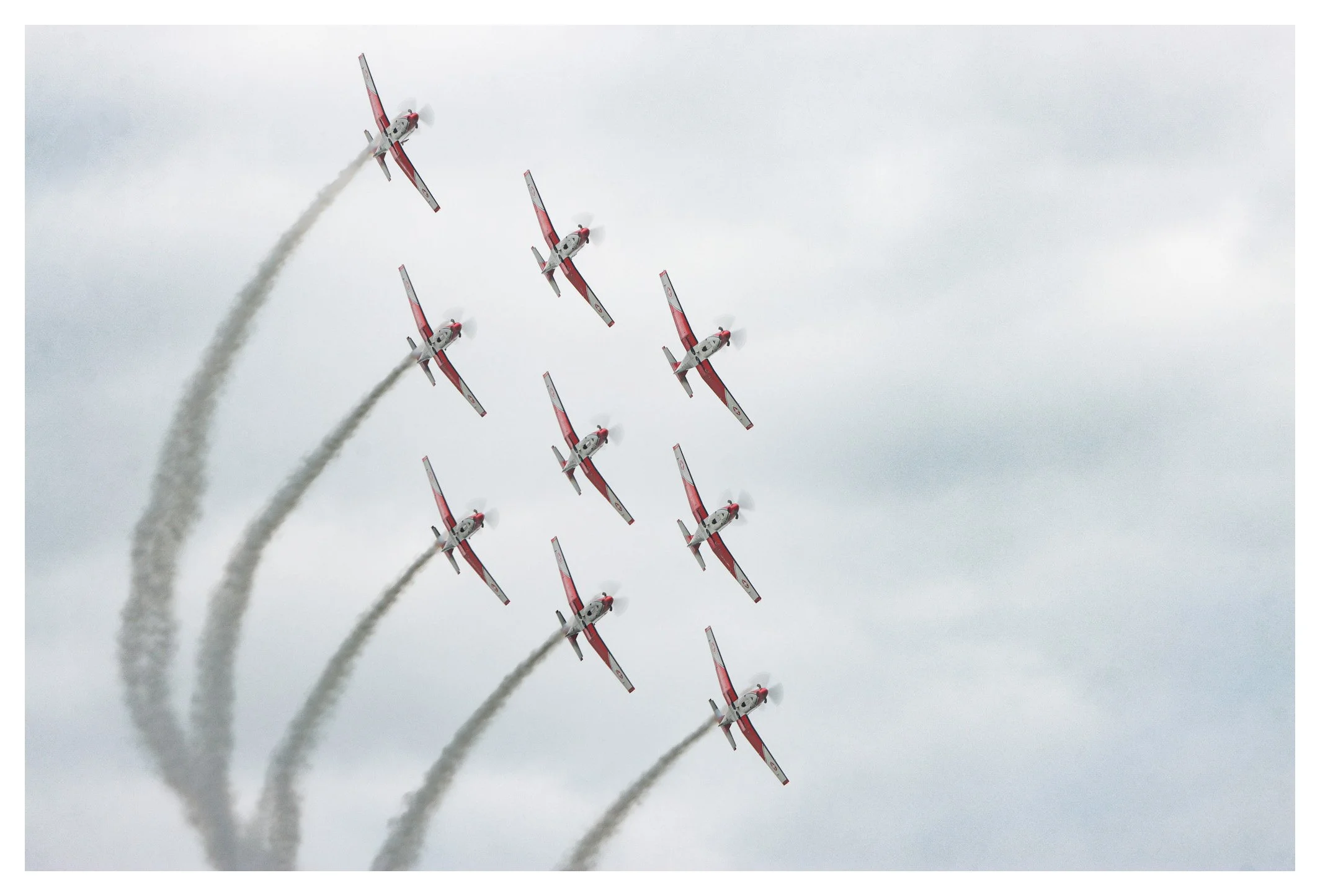 Six red and white airplanes performing an aerobatic formation, each trailing white smoke against an overcast sky.