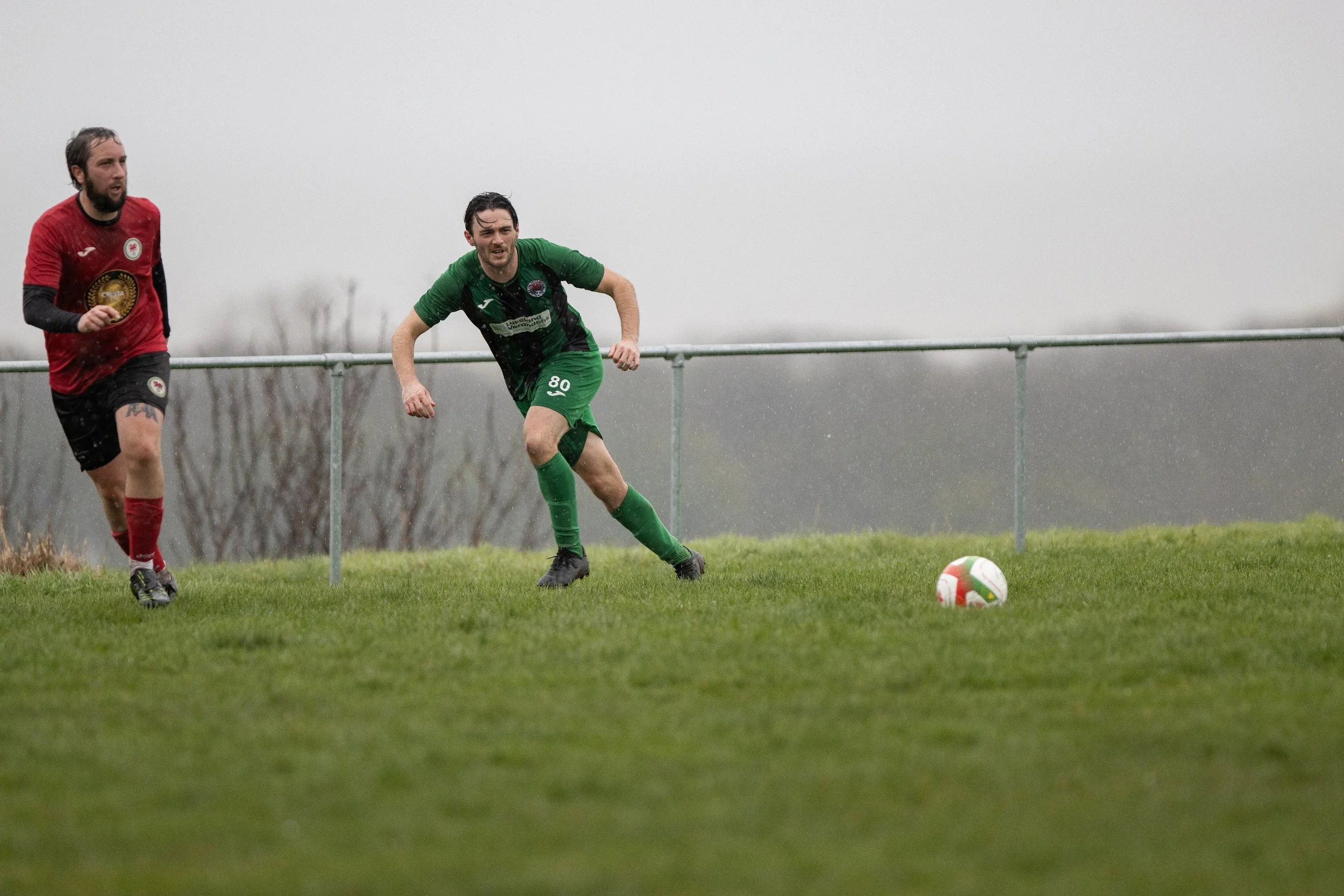 Two soccer players running on a grassy field in rainy weather, one in a red jersey and black shorts and the other in a green and black jersey and green shorts, with a white and red soccer ball nearby.