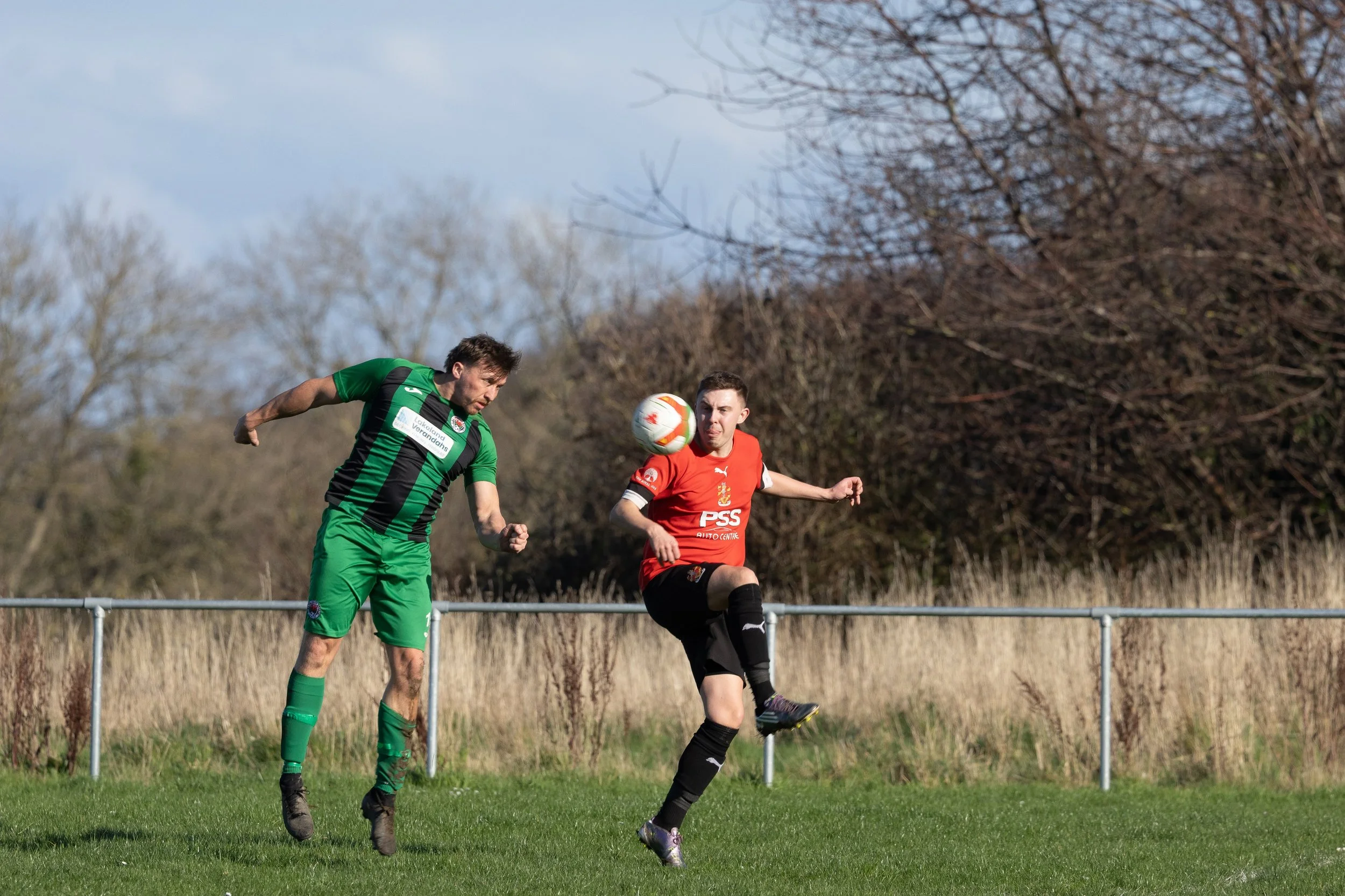 Two soccer players compete for the ball on a grassy field, with trees and a fence in the background.