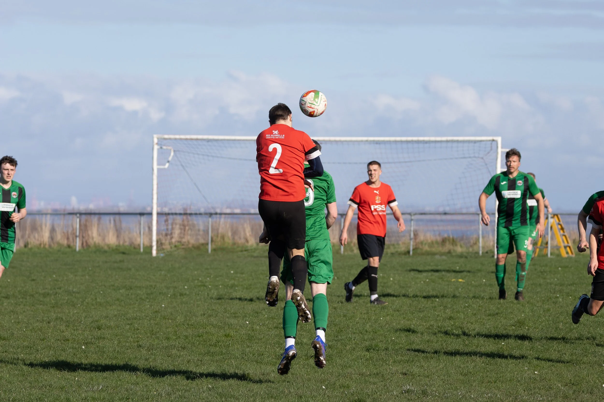 Soccer players on a grass field during a match, with some jumping to head the ball near the goalpost.