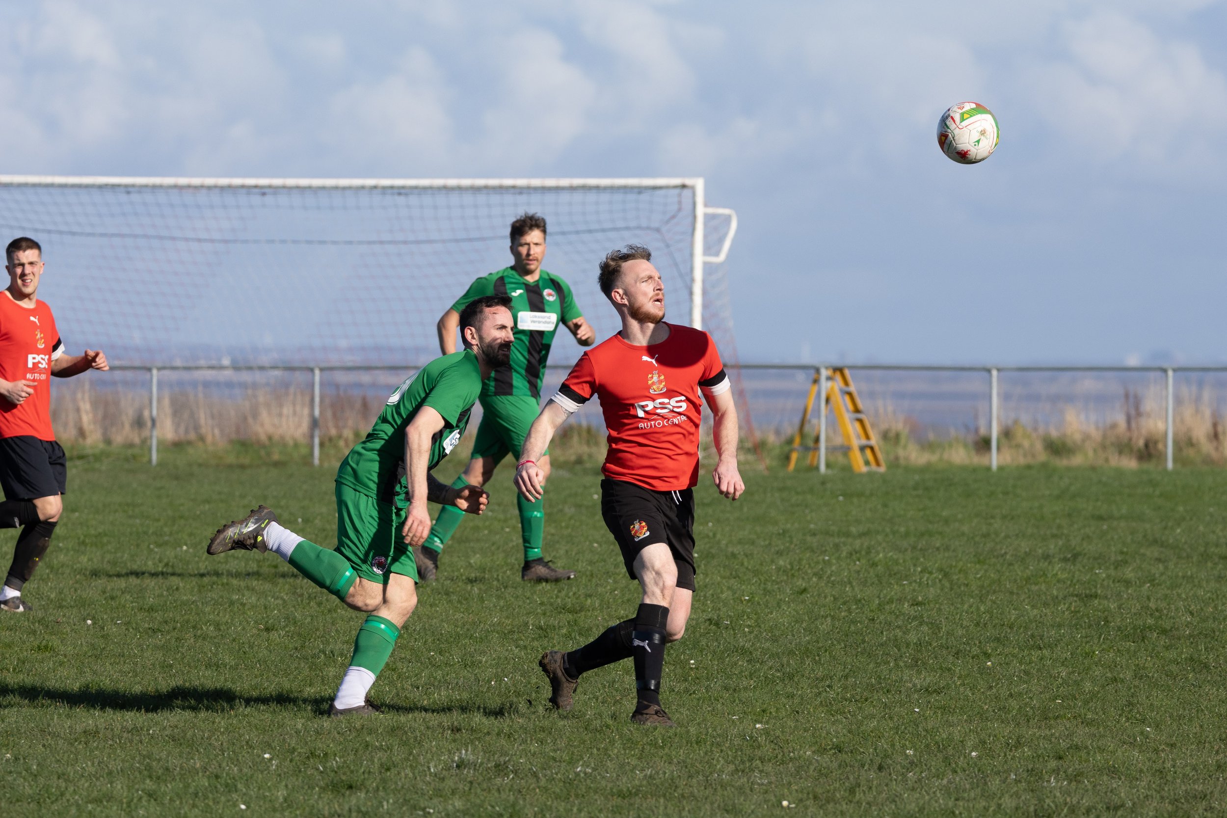 Soccer players on the field during a game. One player in a red jersey head watches the ball in the air, while others in green jerseys are nearby. A soccer goal is in the background.