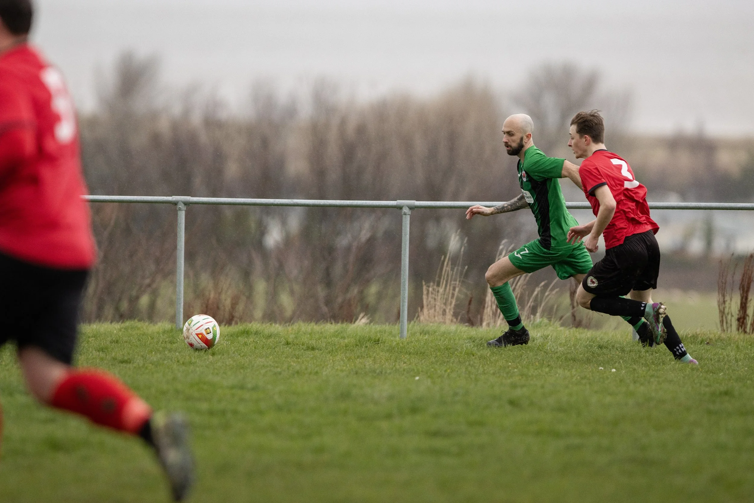 Two soccer players, one in a green jersey and the other in a red jersey, running on a grassy field during a match, with a soccer ball nearby and a blurred background of trees.