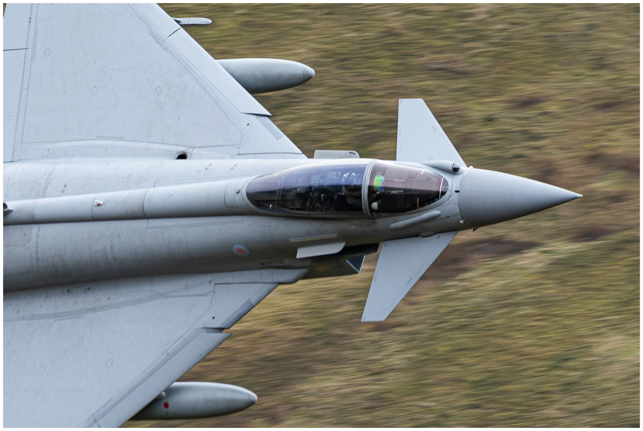 Close-up of a military fighter jet in flight, showing the nose, cockpit, and part of the wing against a blurred background of grass.