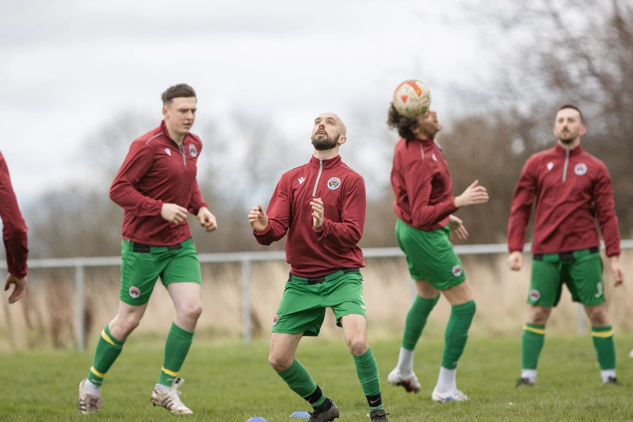 Soccer players warming up on the field, wearing red and green uniforms, with bald and bearded player in the center looking up at the ball