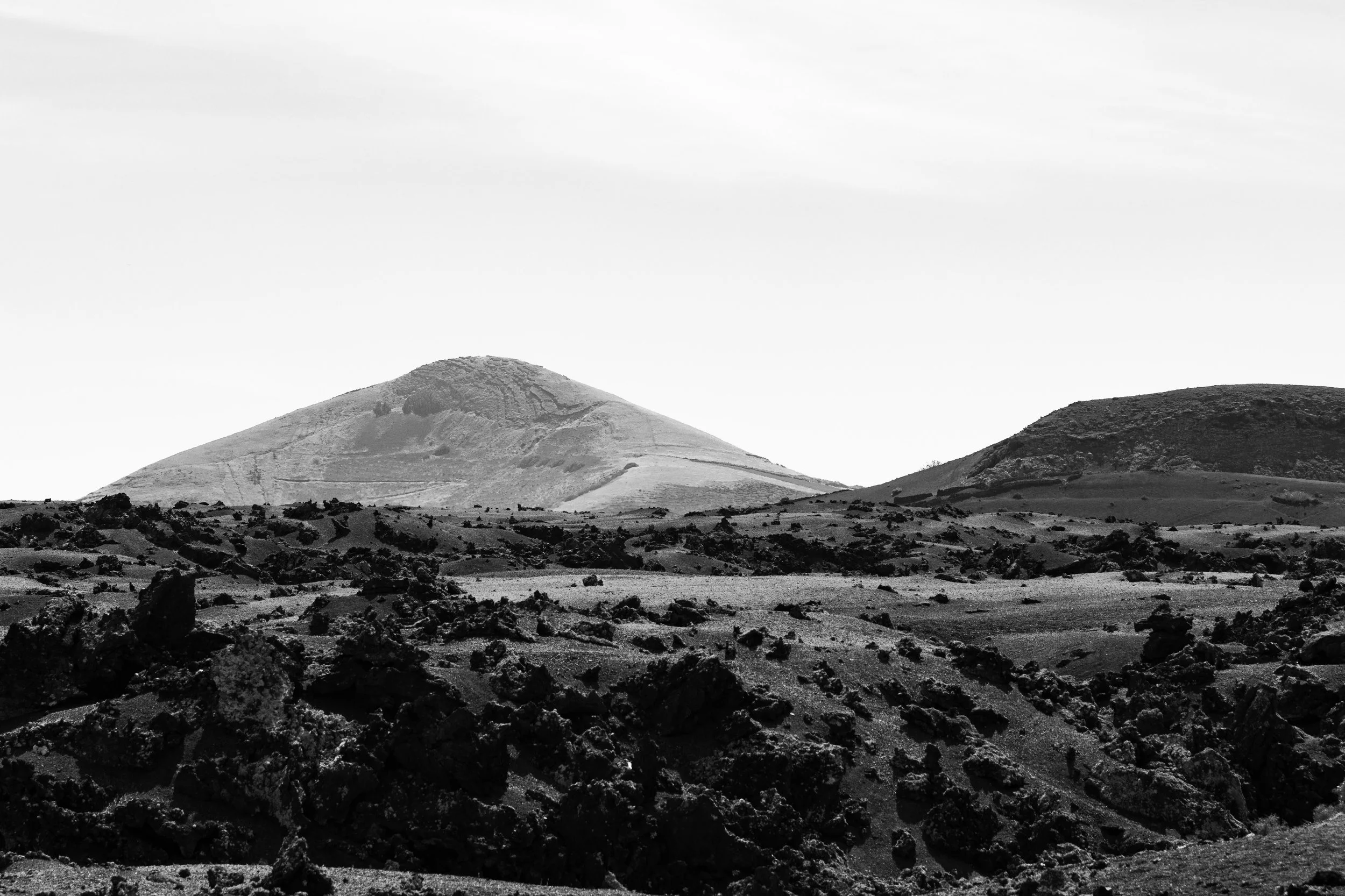 Black and white photo of a rugged volcanic landscape with two volcanic cones in the distance, rough rocks in the foreground, and a mostly cloudy sky.