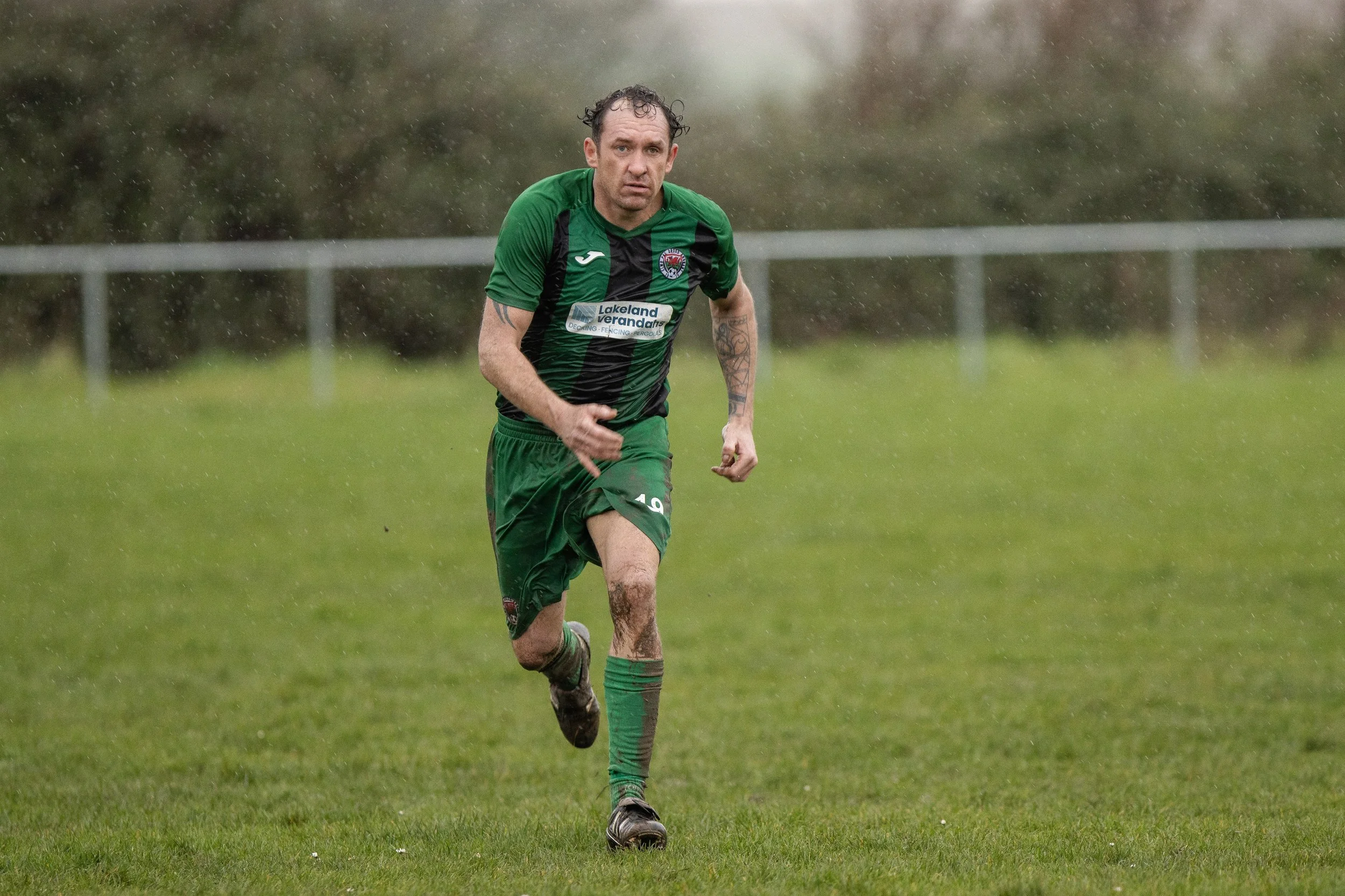 A man running on a wet grassy field while rain is falling, wearing a green and black sports uniform.