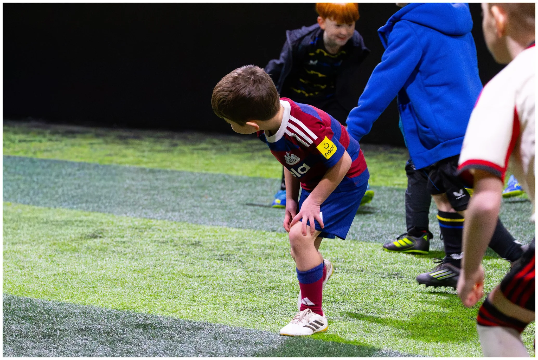 Children on indoor soccer field, some bending over, dressed in sportswear, practicing or stretching.