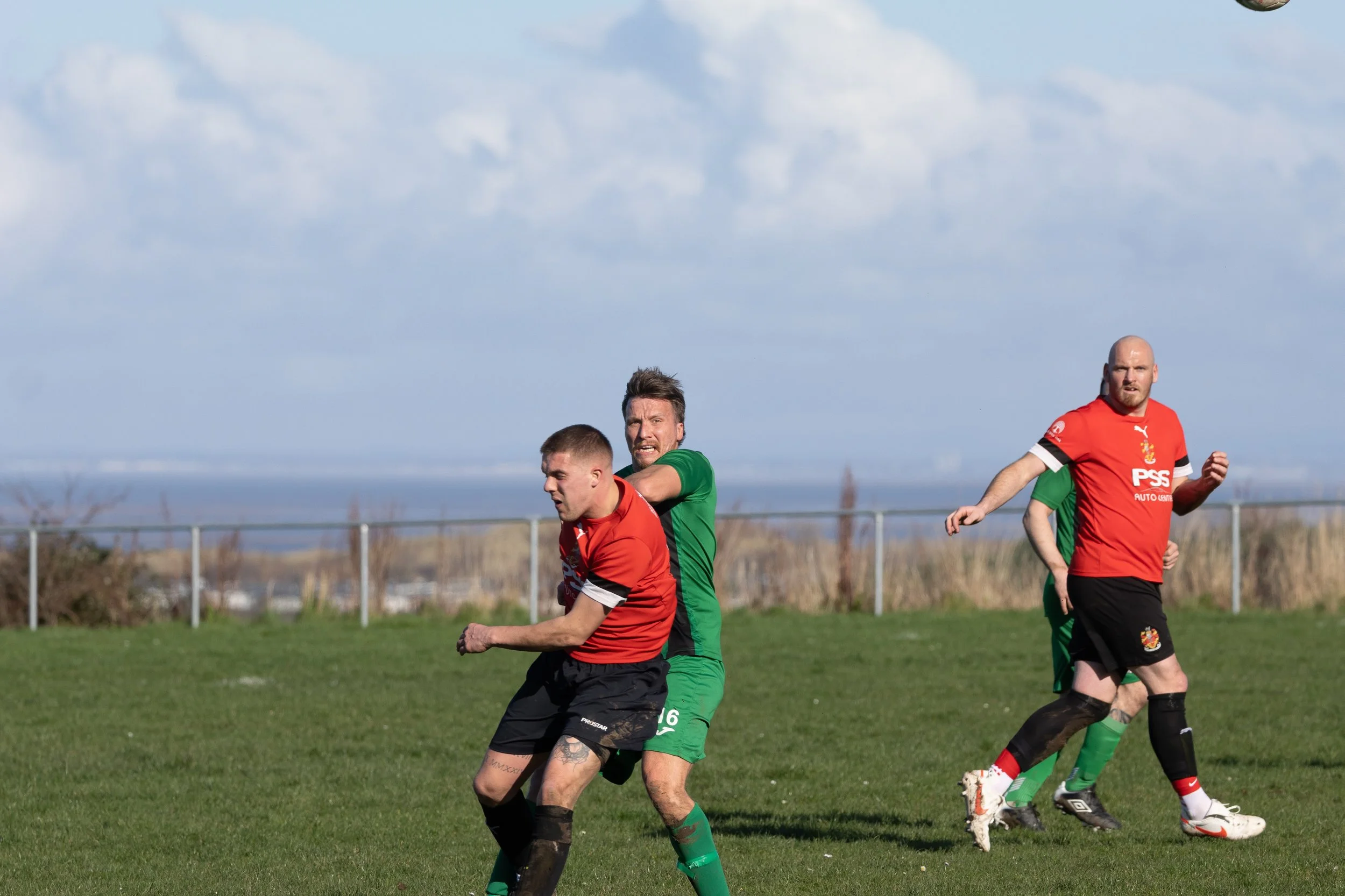 Soccer players in red and green jerseys competing on a grassy field under a partly cloudy sky.