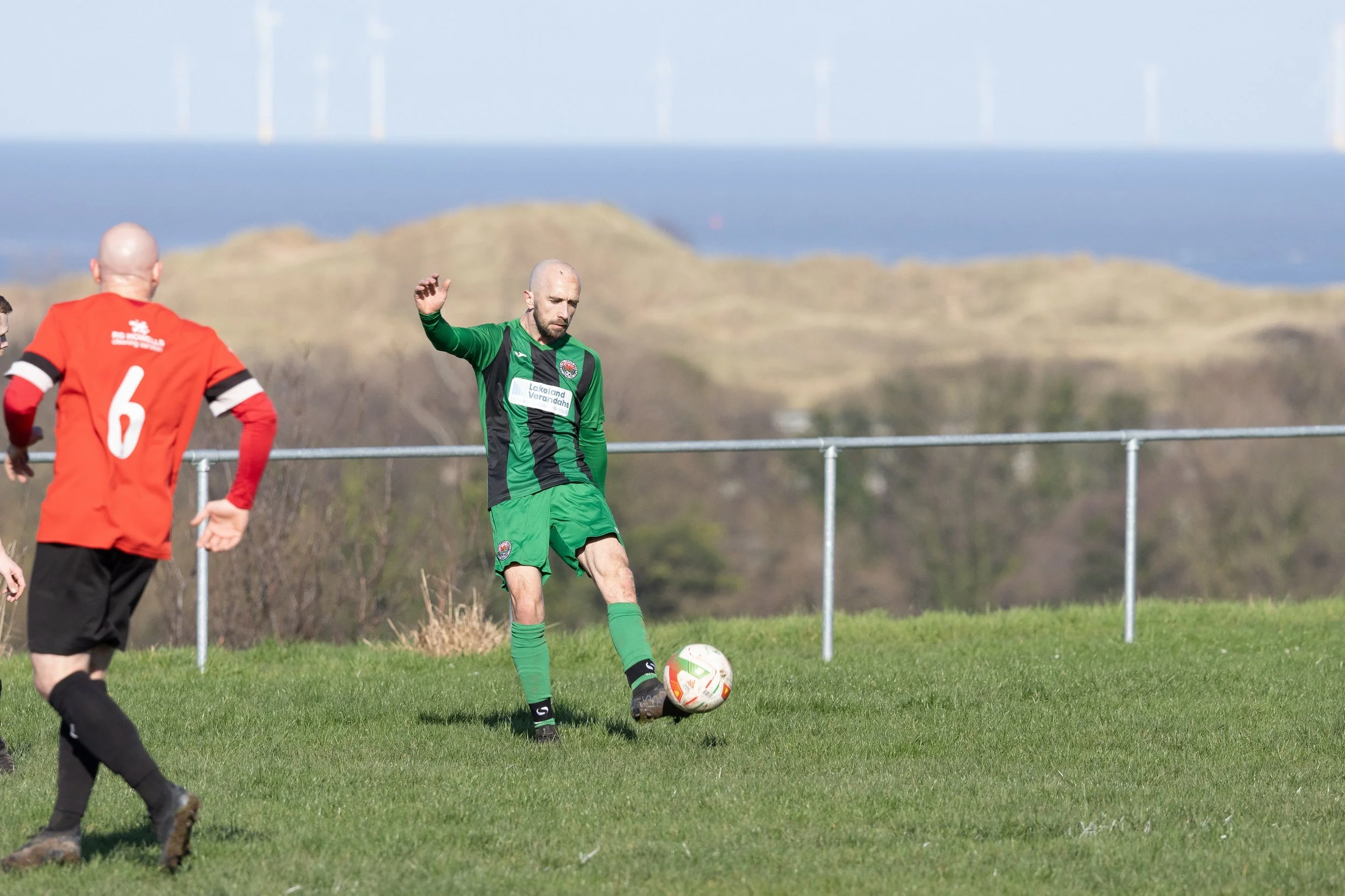 A soccer player in a green and black uniform kicking a ball during a game on a grass field, with an opposing player in a red and black uniform nearby, and a scenic landscape with hills and a wind turbine in the background.