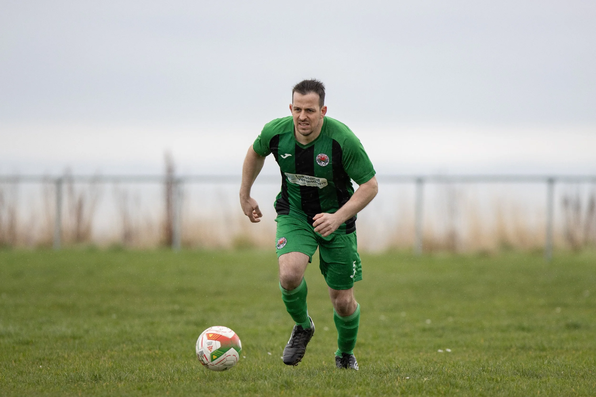 A male soccer player in a green and black uniform running after a soccer ball on a grassy field under a cloudy sky.