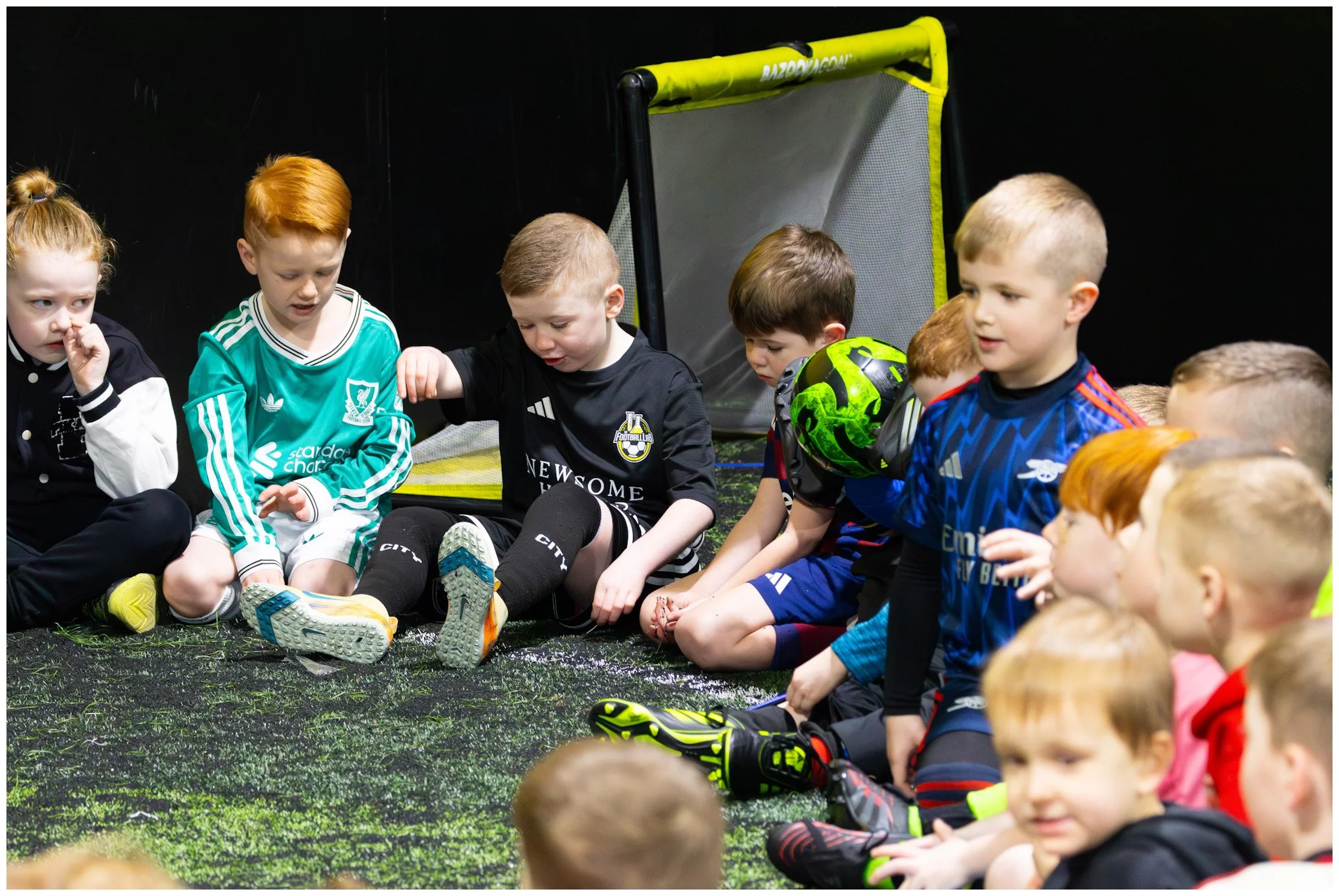 Group of children sitting on the floor, some with soccer gear, inside an indoor sports area.