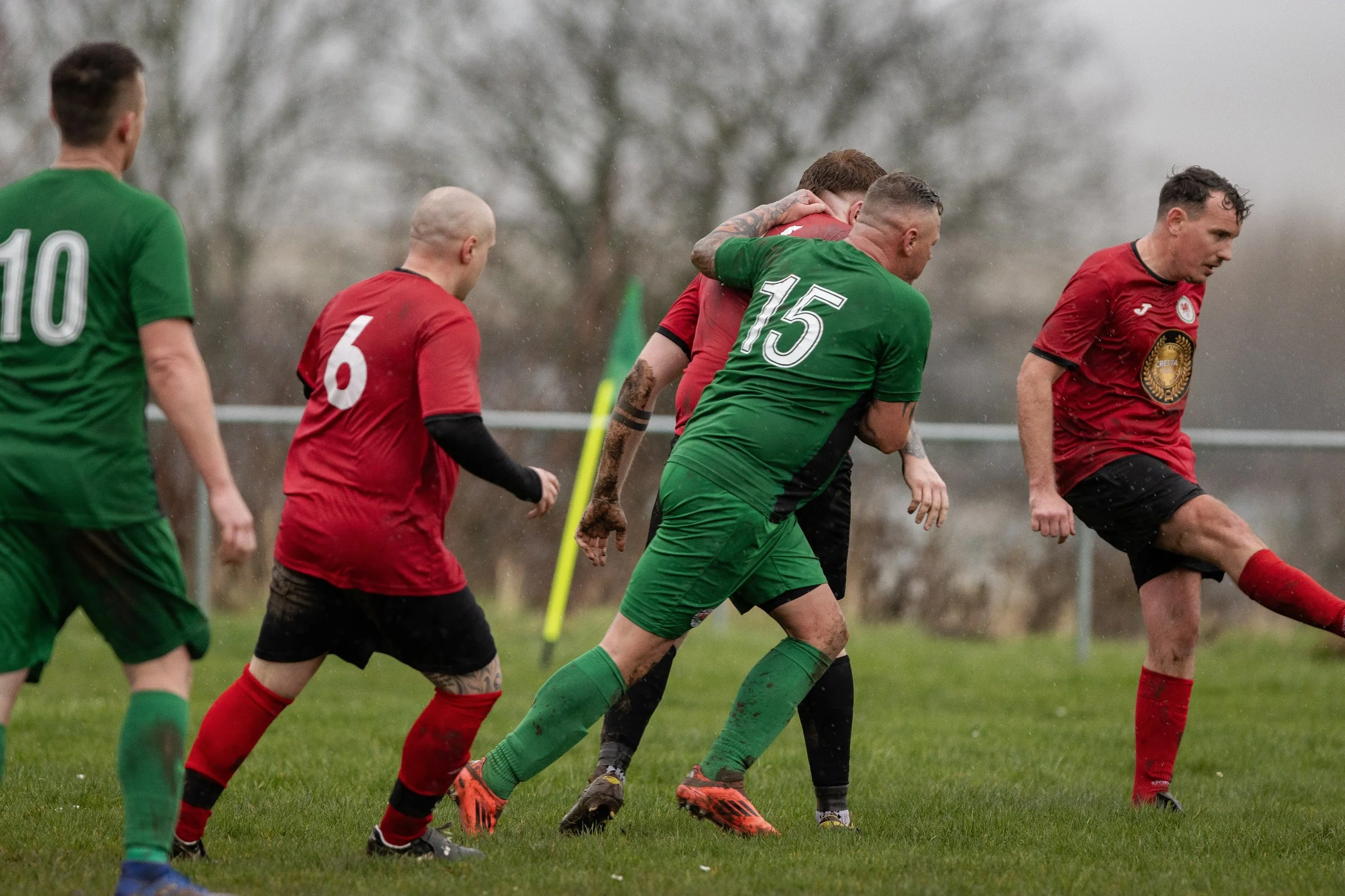 Men playing soccer on a grassy field on a rainy day, with some players wearing green jerseys and others wearing red jerseys, engaged in a close play.
