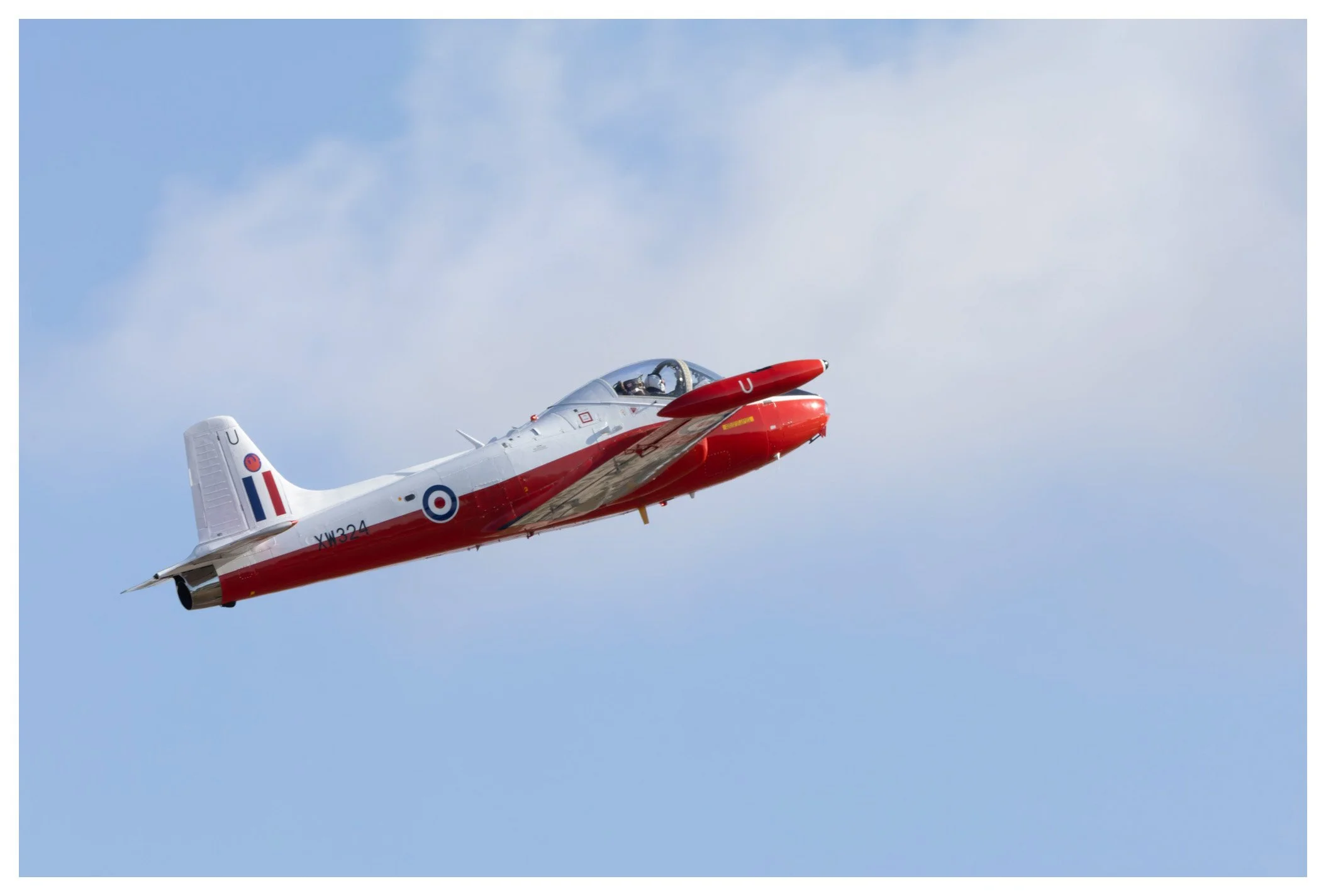 A jet aircraft in red, white, and blue colors flying in the sky with some clouds.