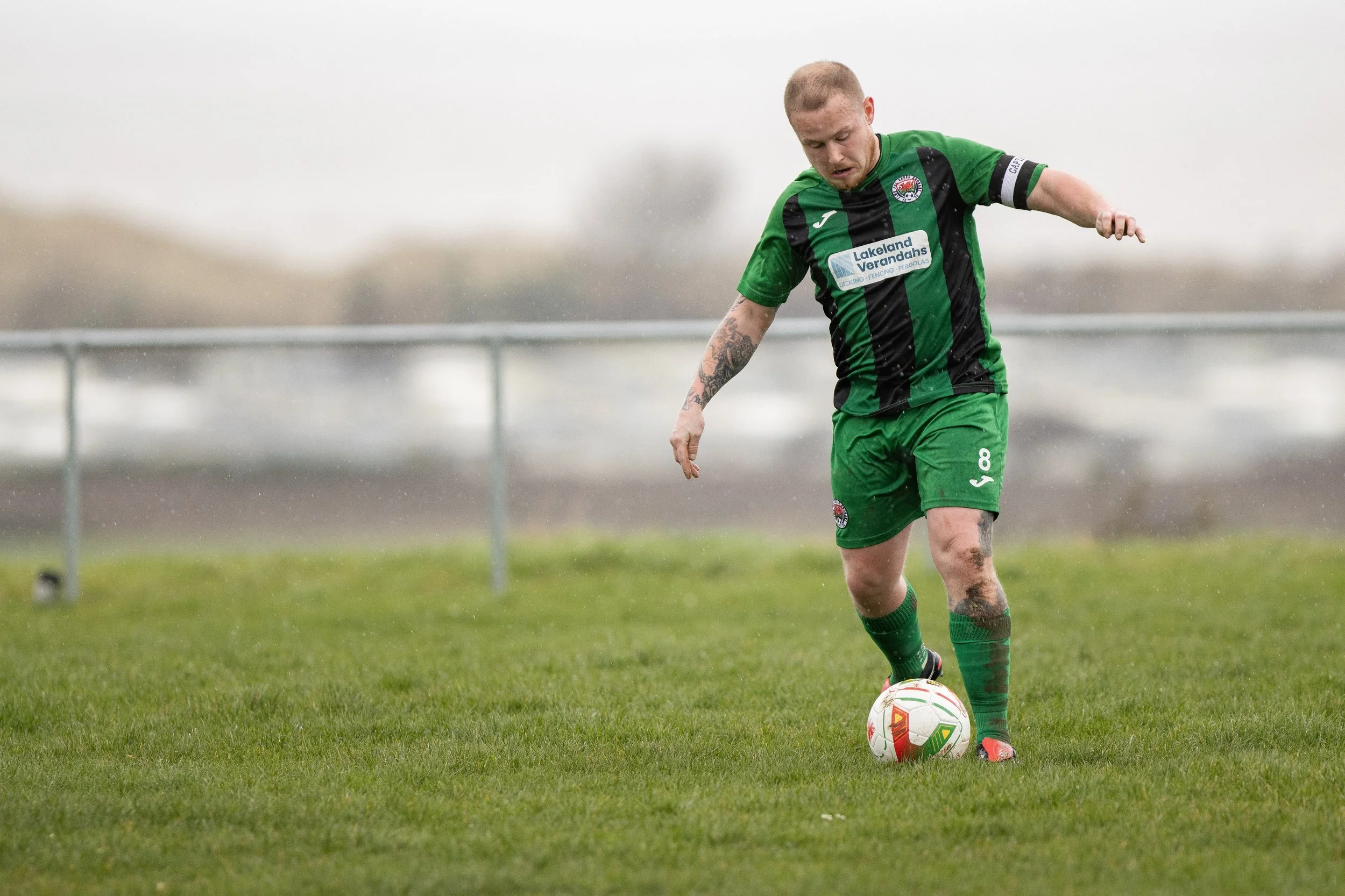 A soccer player in a green and black striped uniform kicking a soccer ball on a grassy field, with a blurred background and overcast sky.