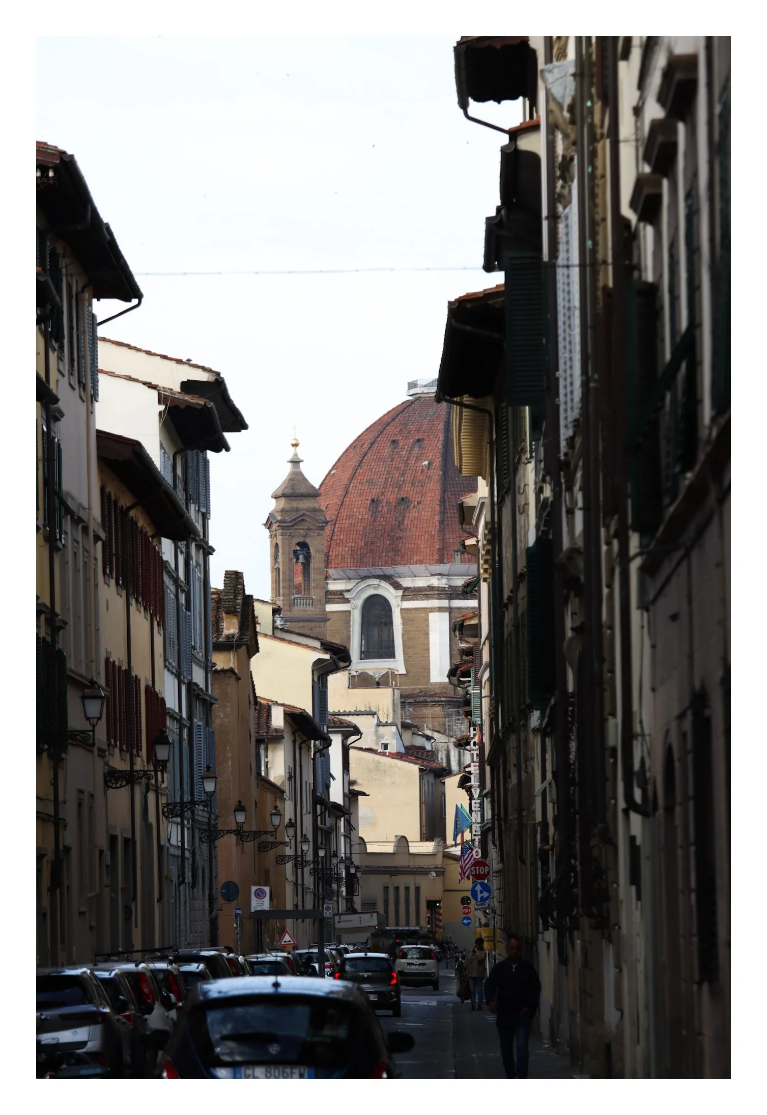 A narrow street with parked cars and pedestrians, lined with historic buildings. In the background, a large domed church with reddish-brown tiles and a small bell tower.
