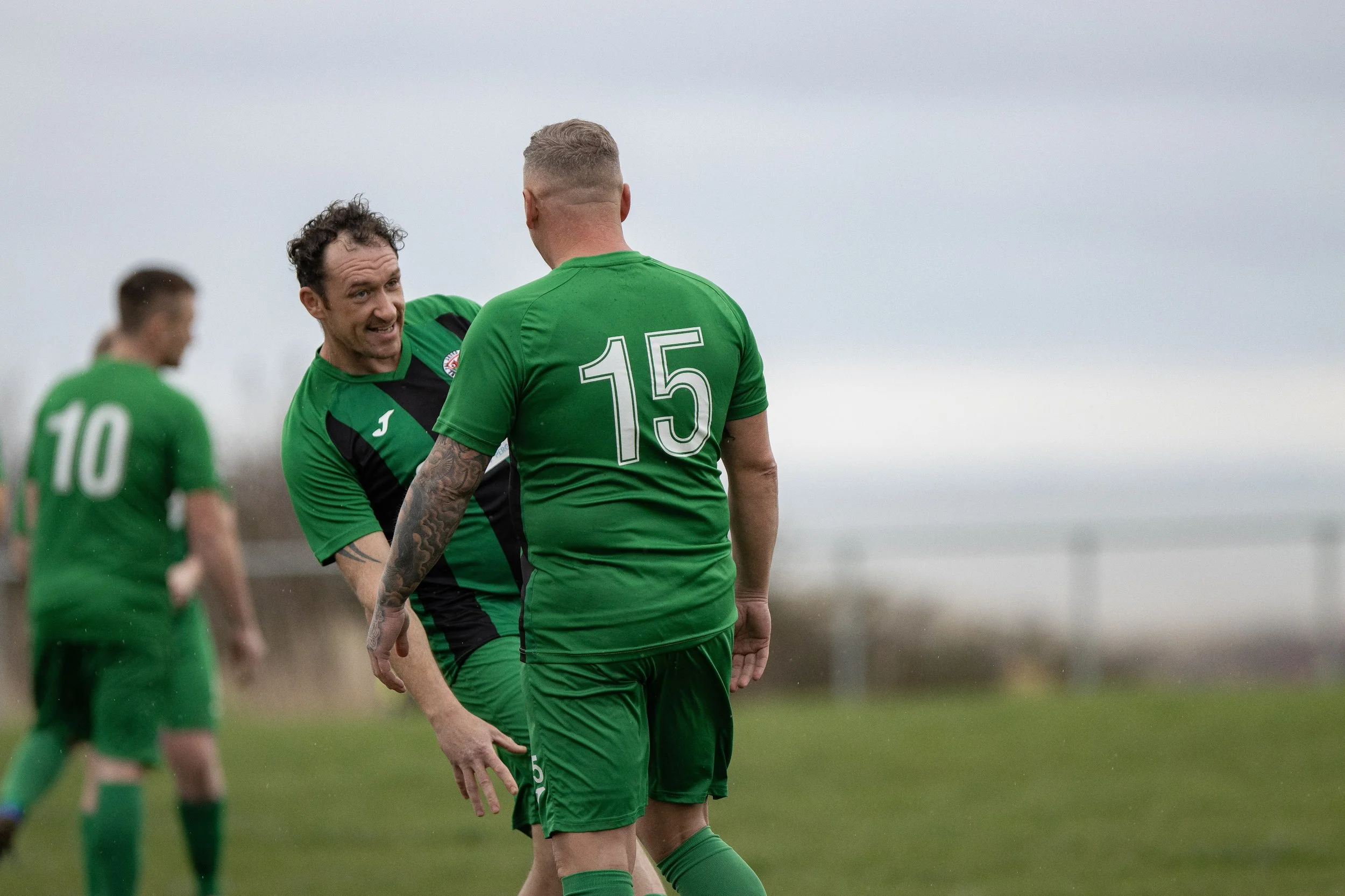 Two soccer players in green jerseys engaging in a friendly interaction on the field, with a third partially blurred player in the background.