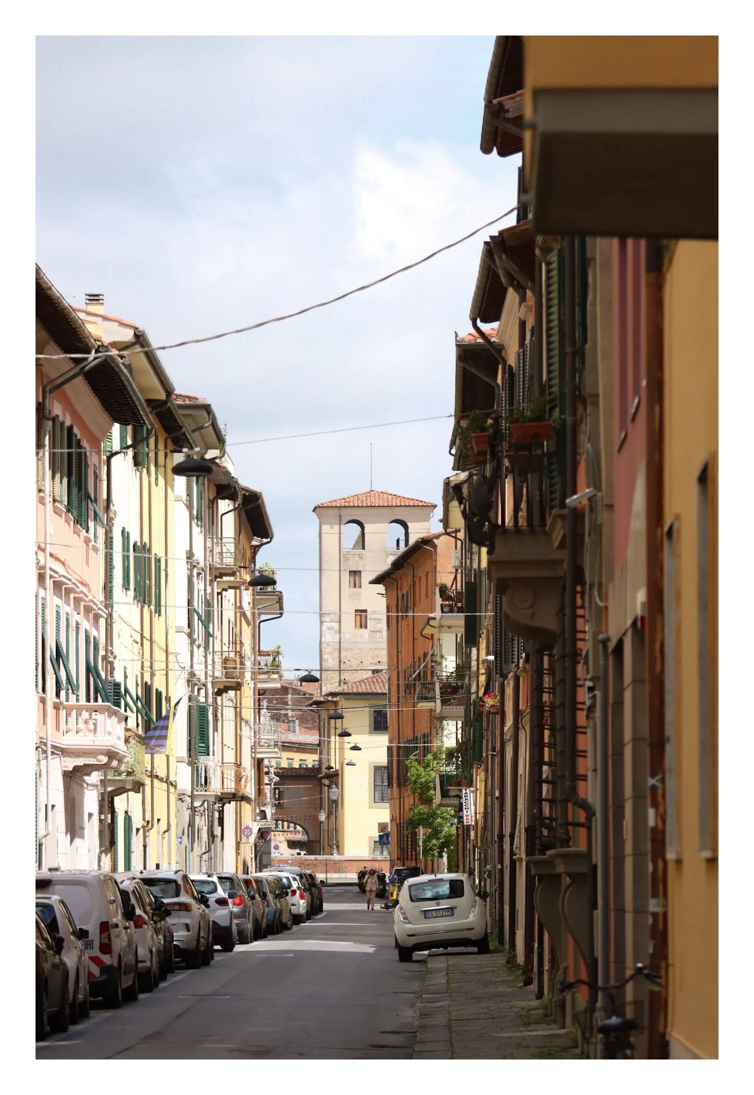 A narrow Italian street lined with colorful buildings, parked cars on each side, and a bell tower with a red-tiled roof in the background.
