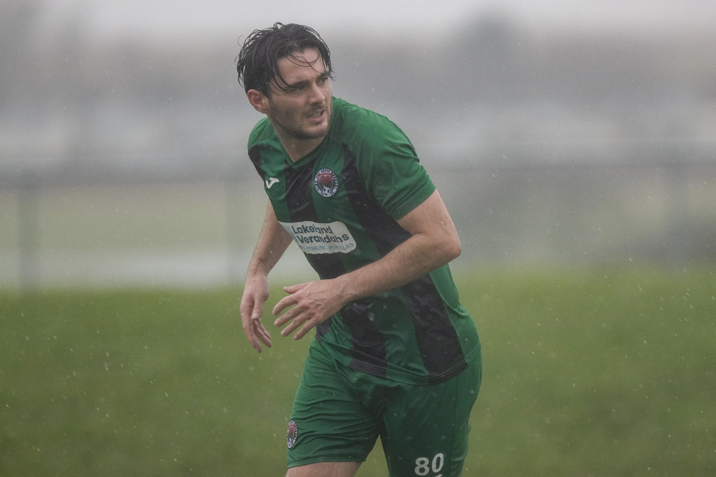 A man in a green and black soccer uniform running in the rain on a grassy field.