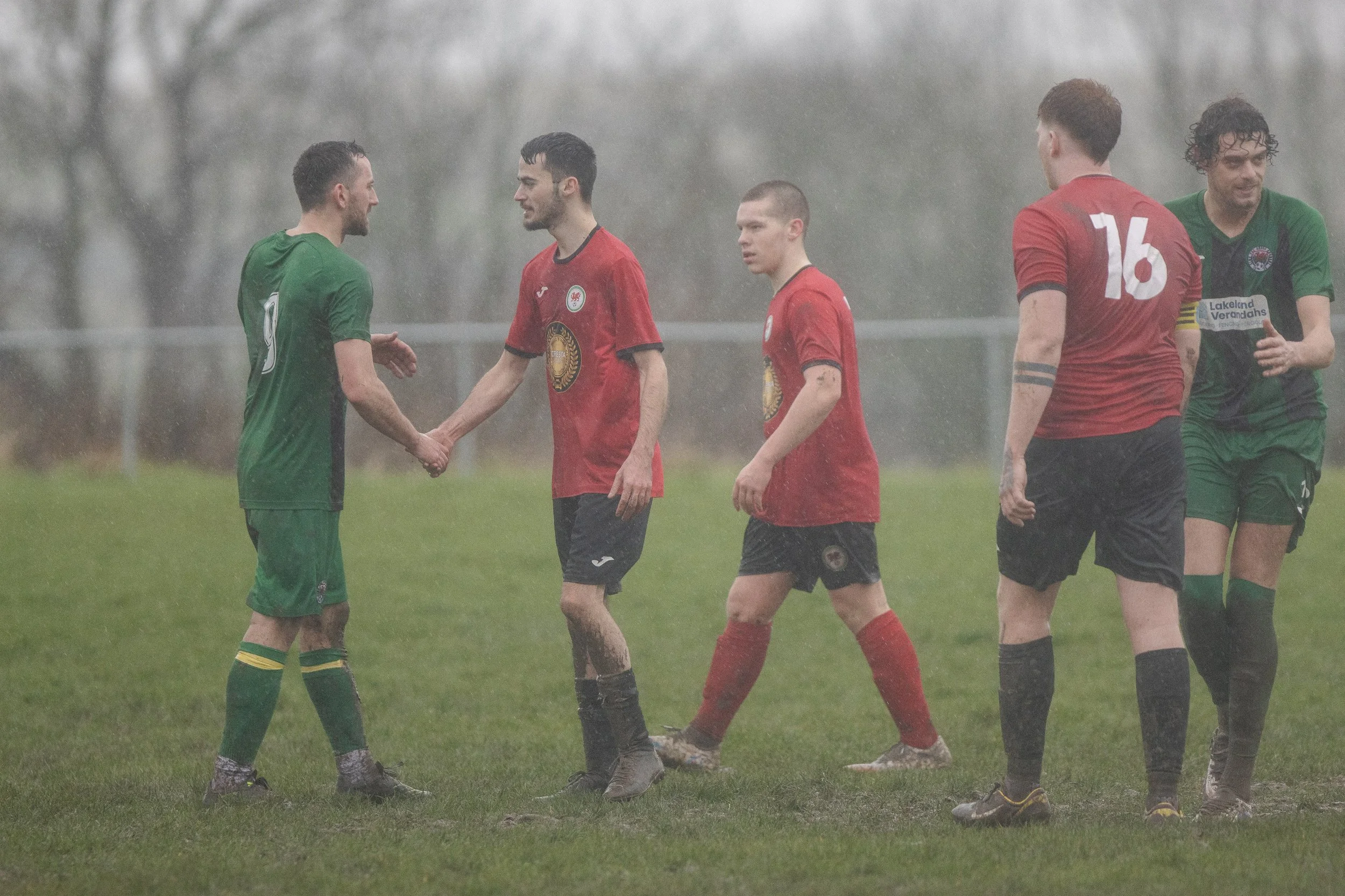 Soccer players shaking hands on a rainy field, wearing red and green uniforms.
