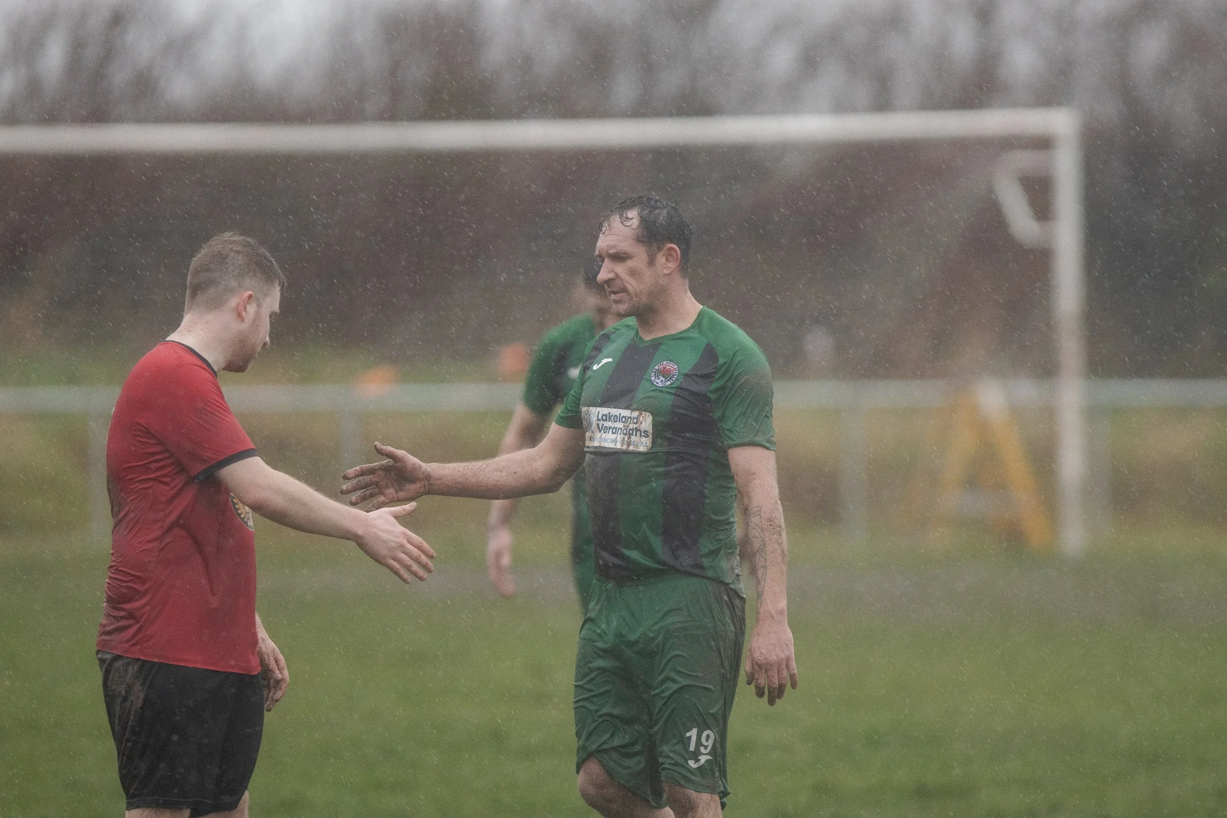 Two soccer players shaking hands in the rain on a soccer field. One player is wearing a red jersey, and the other is in a green and black jersey.