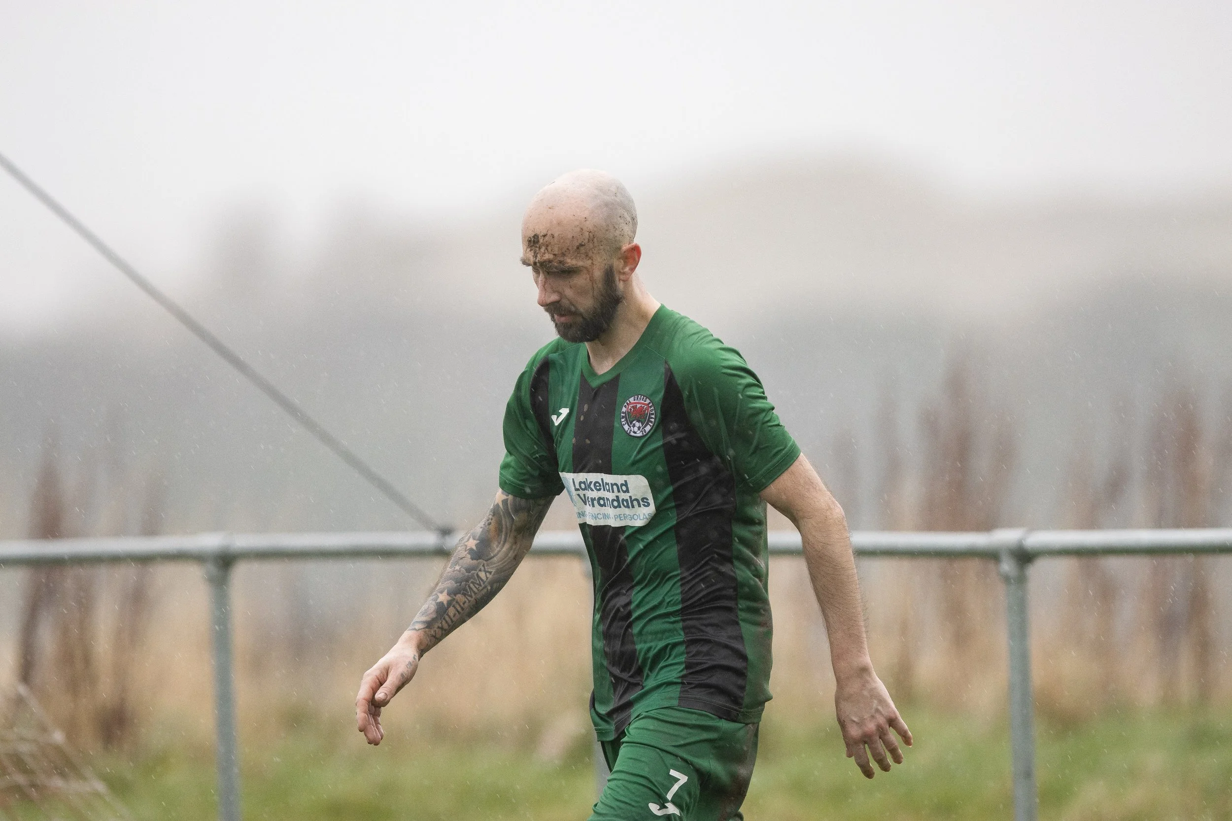 A bald male soccer player with tattoos on his left arm, wearing a green and black jersey, walking on a field in rainy weather with a somber expression.