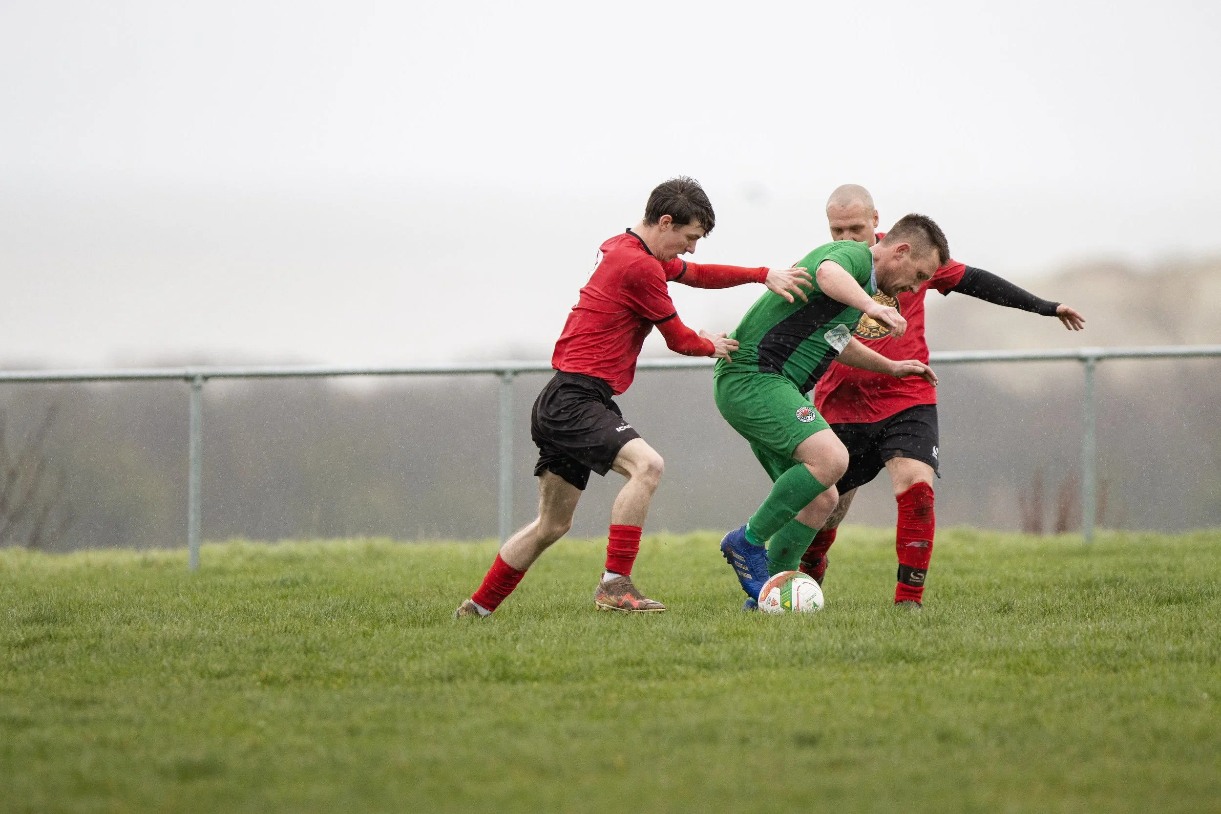 Three soccer players in red and green jerseys compete for the ball on a grassy field, with two players in red and one in green.