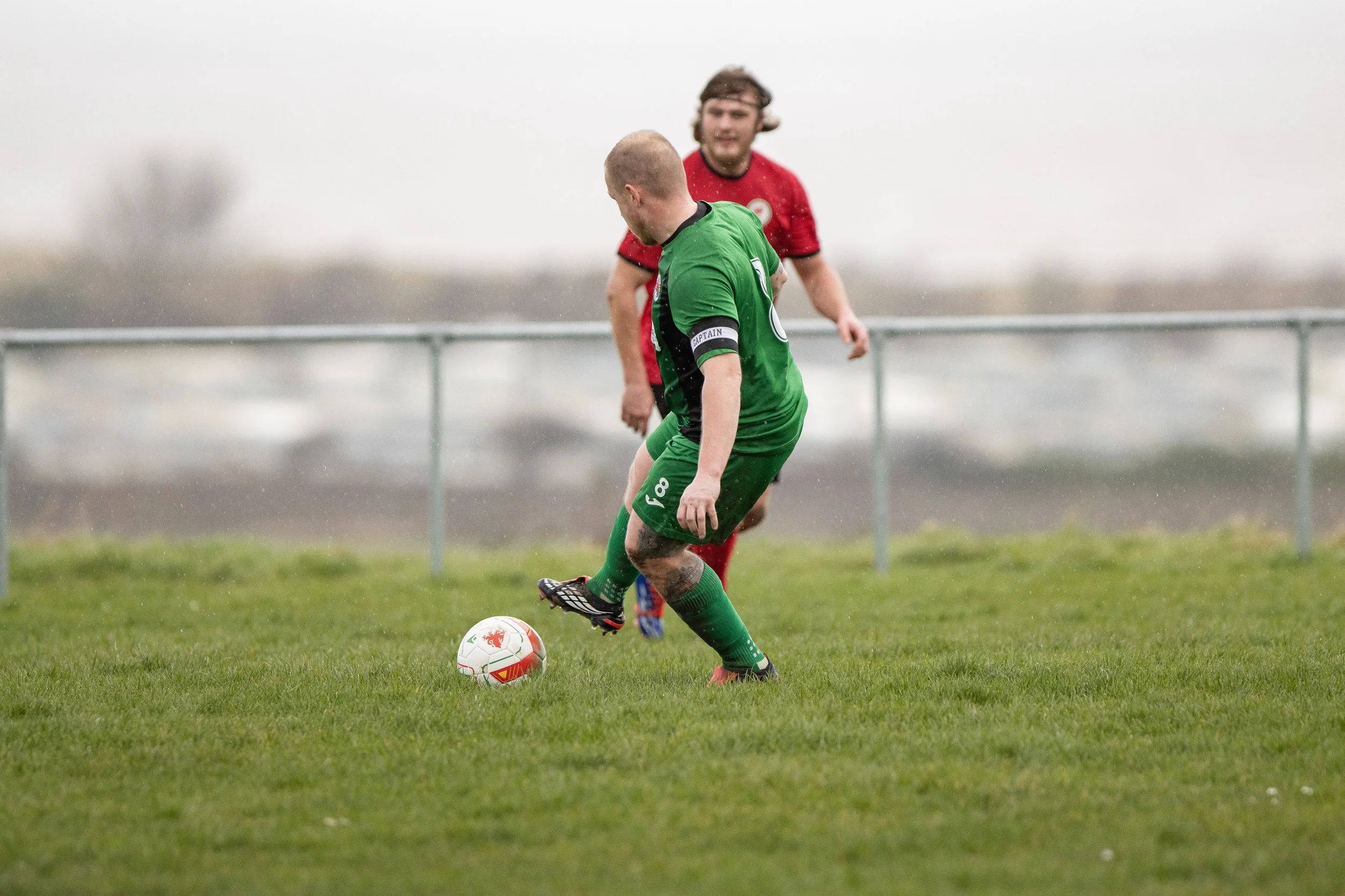 Two soccer players in green and red uniforms compete for the ball on a grassy field during a rainy day.