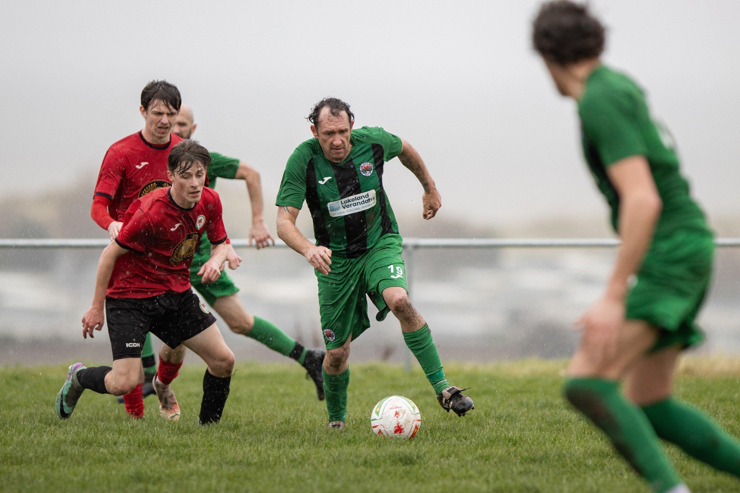 A group of people playing soccer on a rainy day on a grassy field, with some players wearing red jerseys and others in green, chasing the ball.