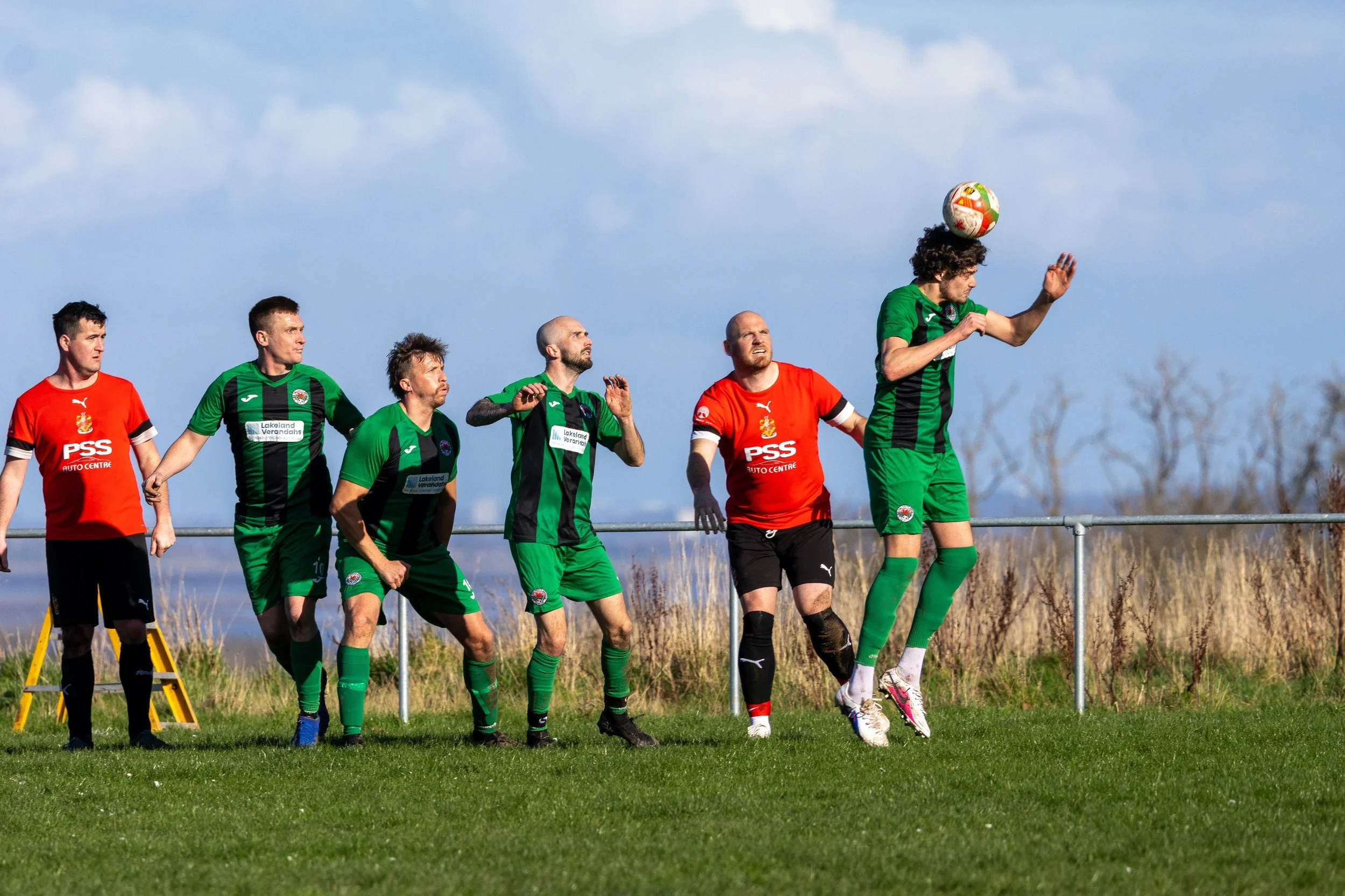 Soccer players in green and red uniforms jumping and running to head the ball during a match on a grassy field with a blue sky background.