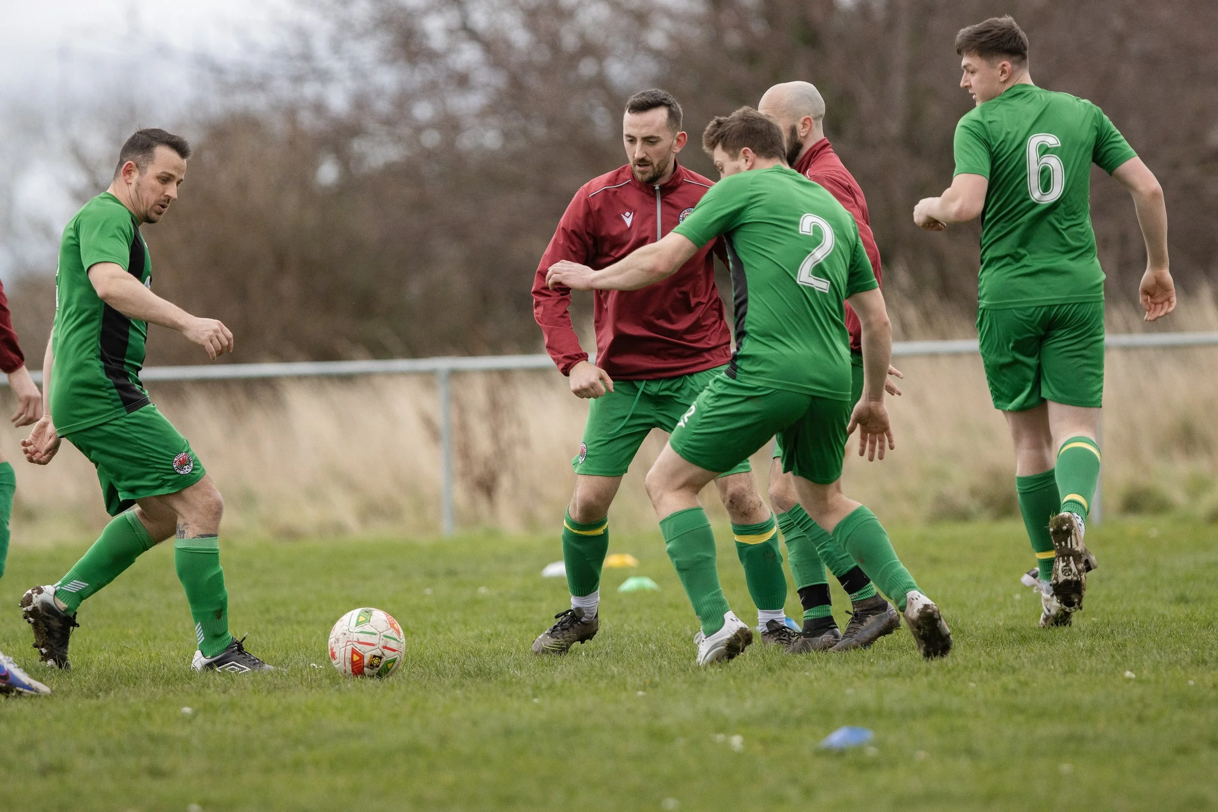 A group of male soccer players on the field during a game, wearing green and maroon jerseys, with some players trying to gain control of the soccer ball.